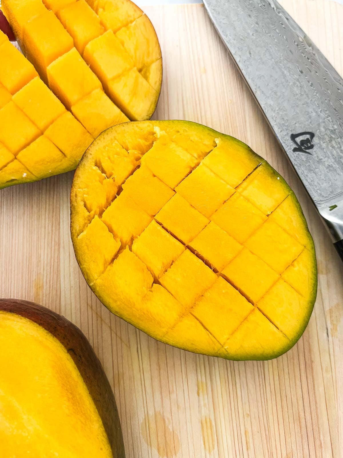 open half of a mango with a cross-hatch score, showing how to slice a mango - on a wood cutting board next to a knife.