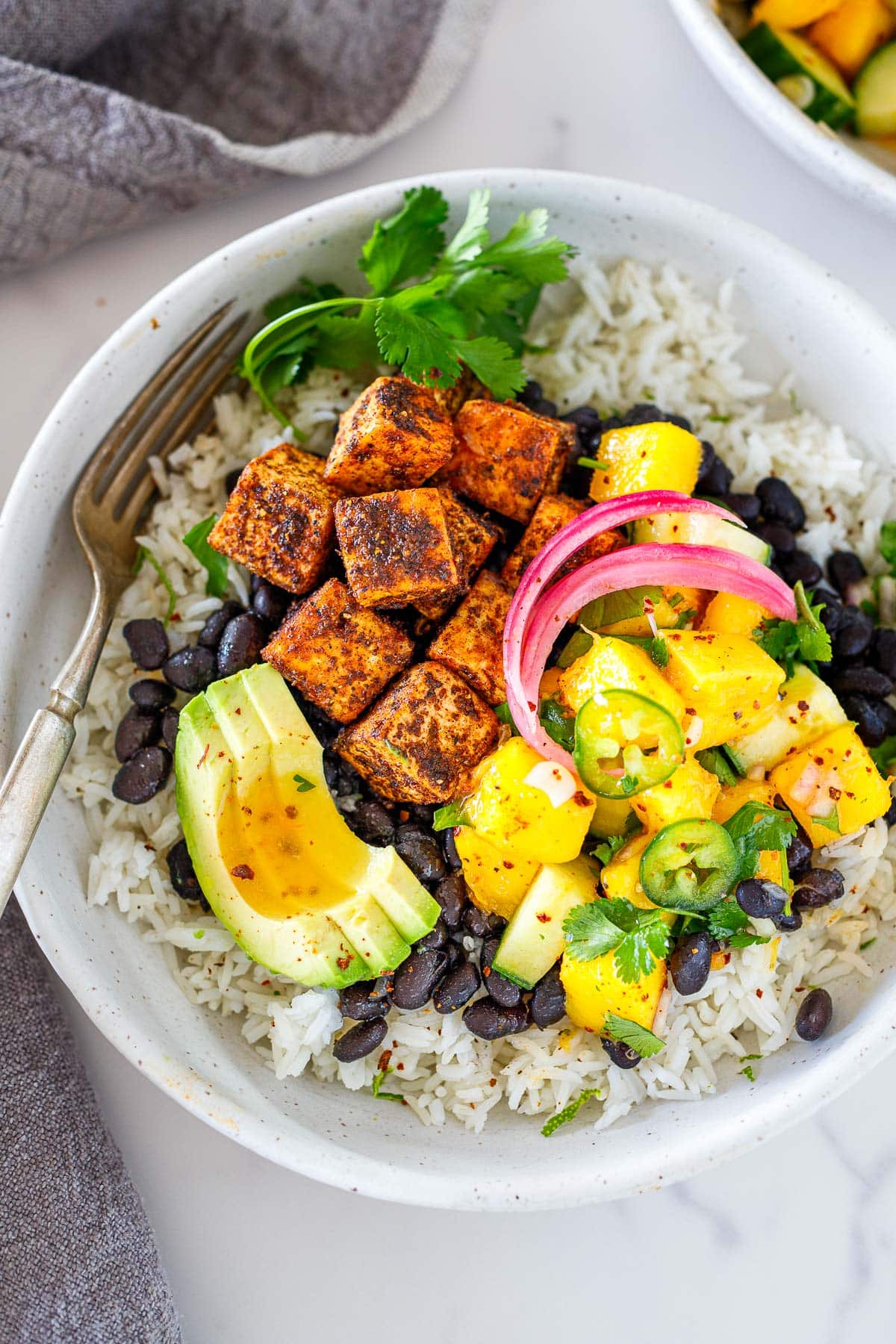 spicy jerk tofu bowl with mango salad, pickled onion, and sliced avocado on bed of rice and black beans in bowl with fork.
