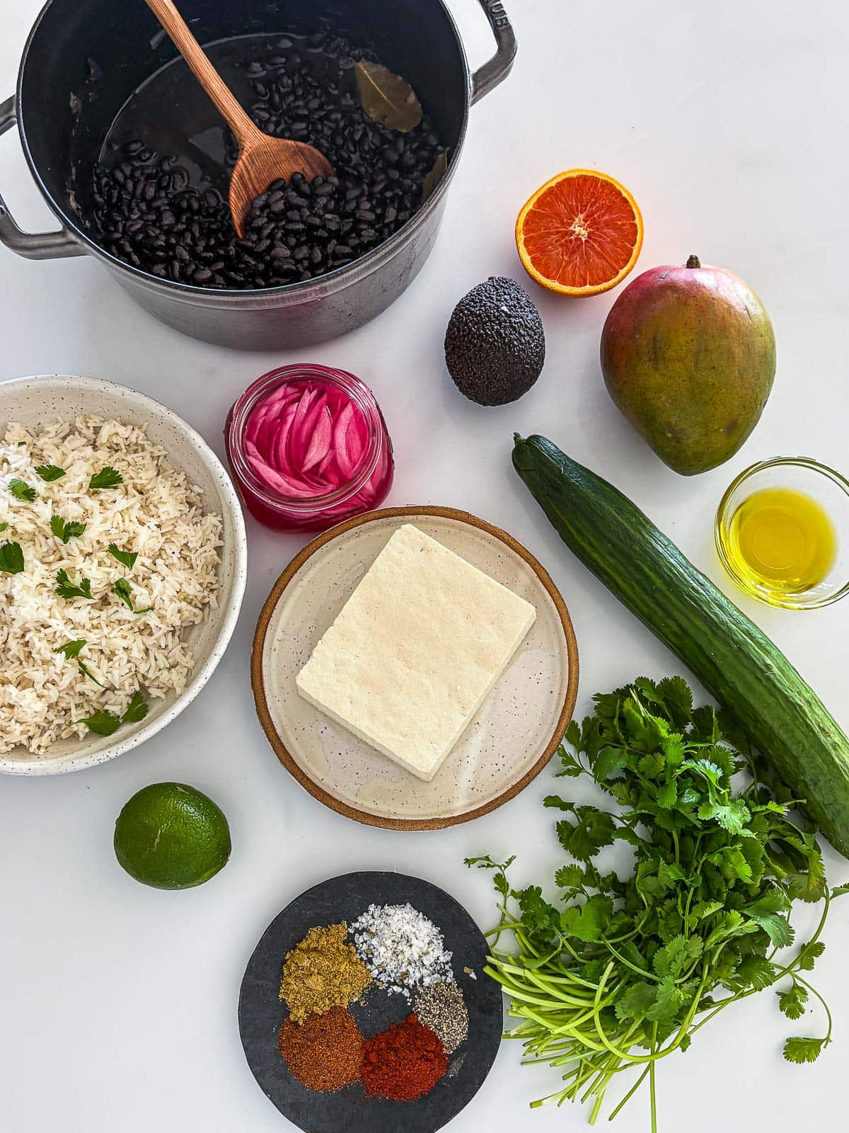 ingredients laid out for tofu bowl - pot of cooked black beans, orange, mango, avocado, oil, cucumber, cilantro, tofu, spices, lime, pickled onions, and rice.