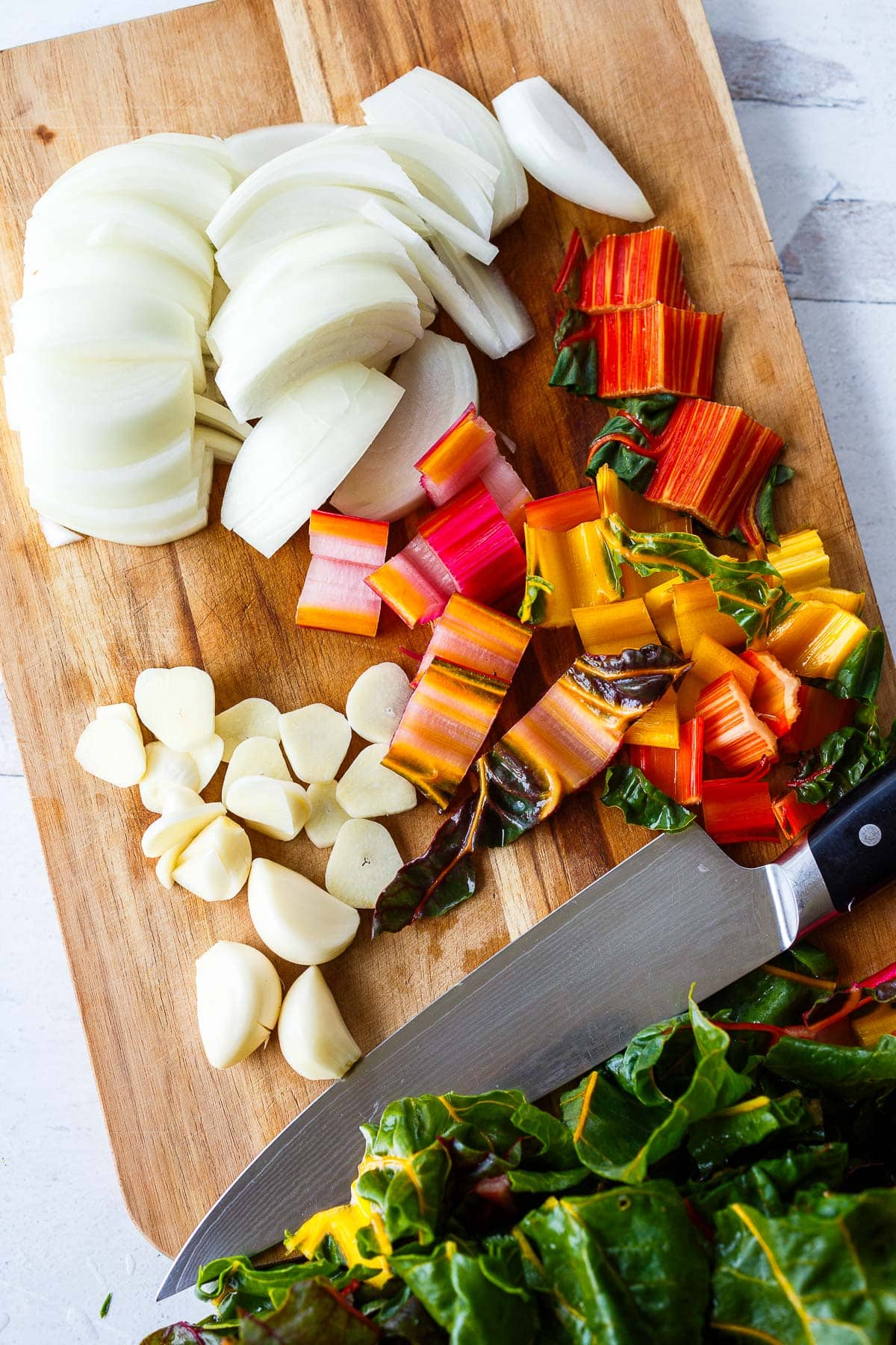wood cutting board, sharp knife, and sliced and chopped veggies - onion, chard stems, and garlic.