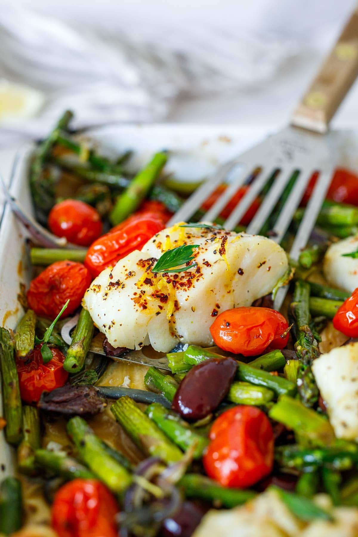 metal spatula lifting up baked white fish seasoned with red pepper flakes, lemon zest, and parsley on pan with olives, tomatoes, and asparagus.