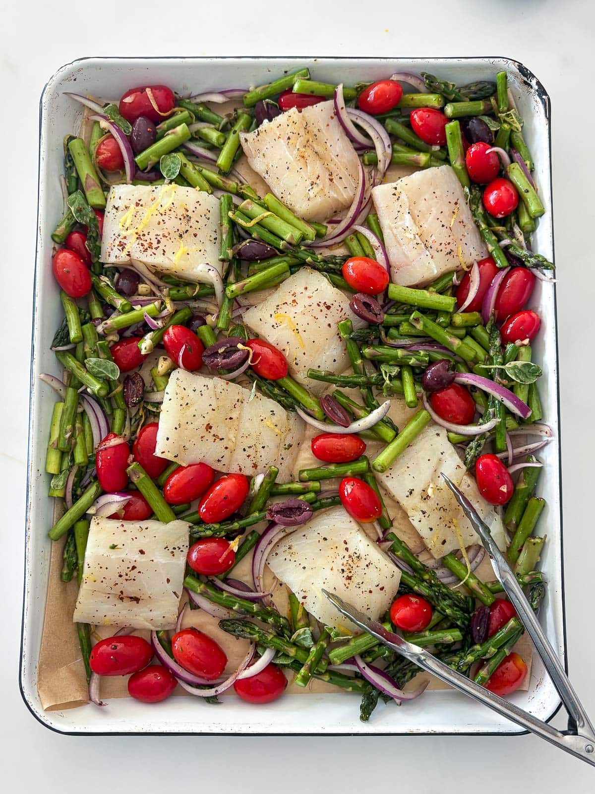 baking sheet with cod seasoned with pepper, red pepper flakes, and lemon zest, tucked between asparagus, cherry tomatoes, olives, and red onion.