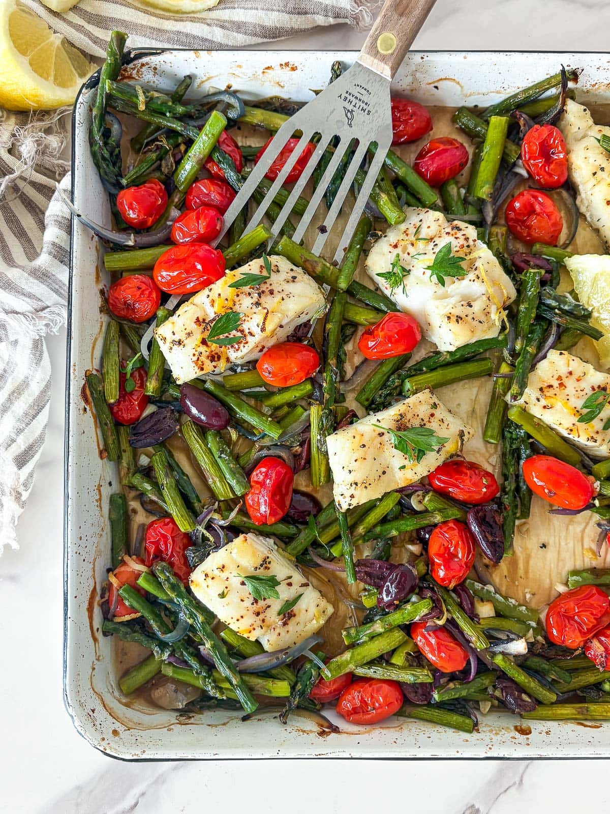 baking sheet with parchment with Mediterranean baked cod nestled between roasted asparagus, burst cherry tomatoes, olives, and red onions, with metal spatula scooping up the fish.