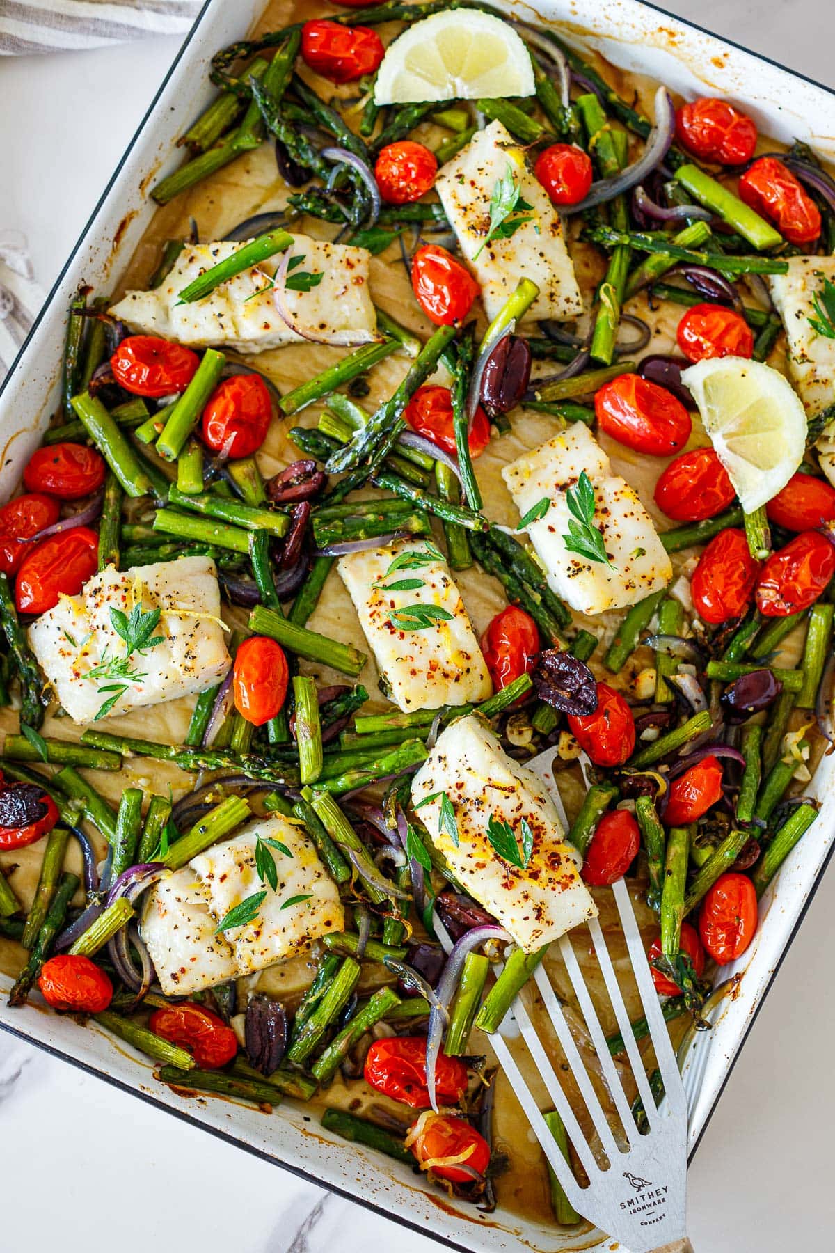 baking sheet with parchment with Mediterranean baked cod, cherry tomatoes, asparagus, olives, and red onion.