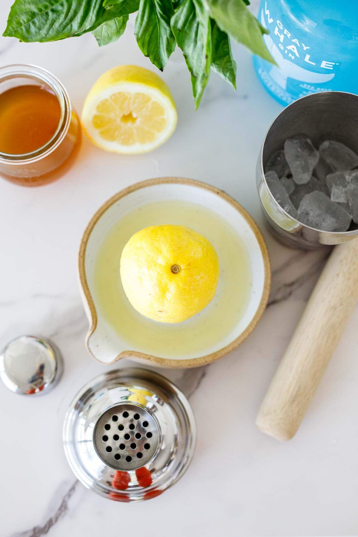 ingredients arranged to make a basil variation of the bee's knees - gray whale gin, shaker with ice, fresh basil, honey syrup, lemon half, lemon in juicer plate, muddler, and shaker lid.