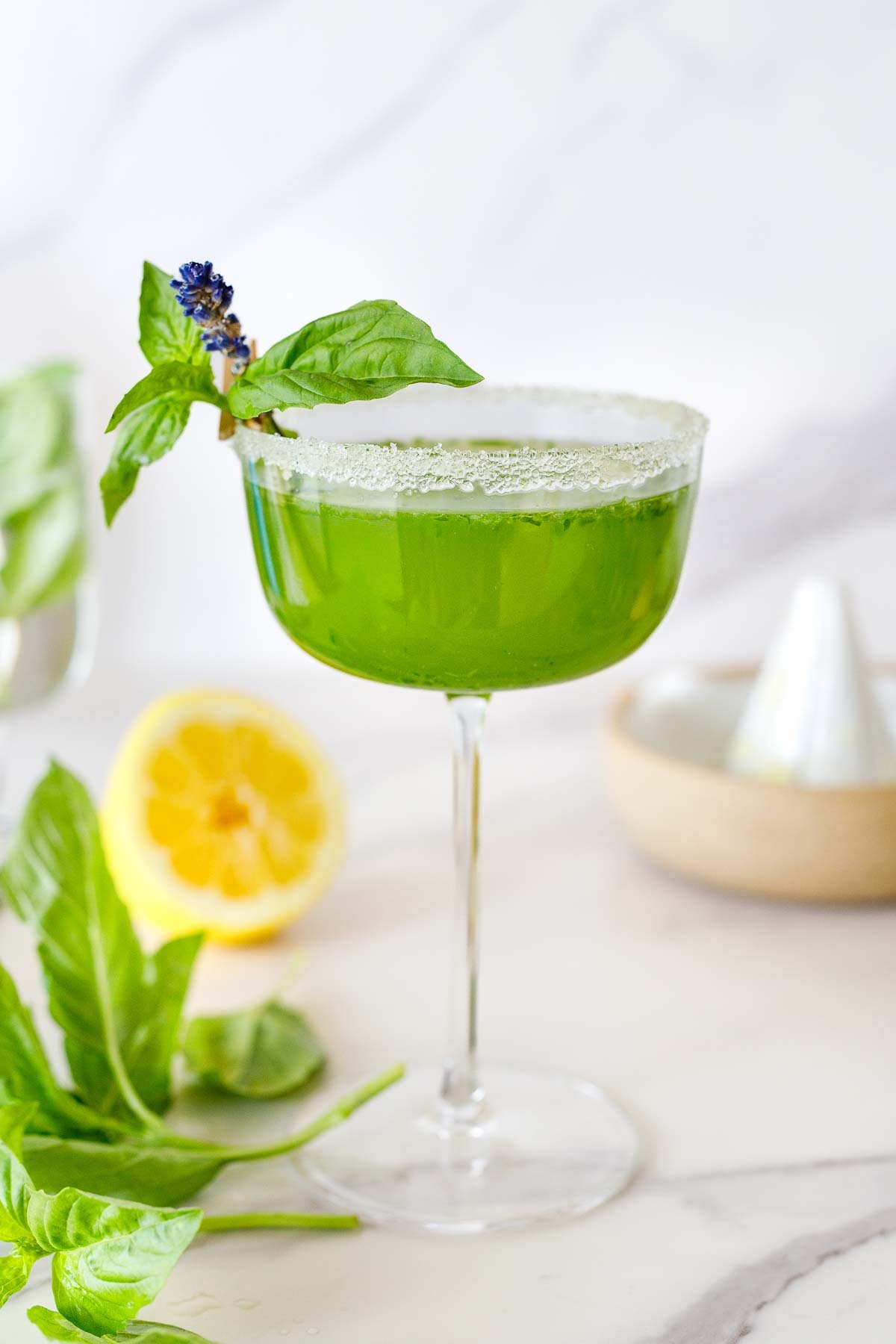 basil bee cocktail in a coupe glass, a basil variation of the bee's knees, garnished with a sugar rim and basil and lavender attached with a clothespin.