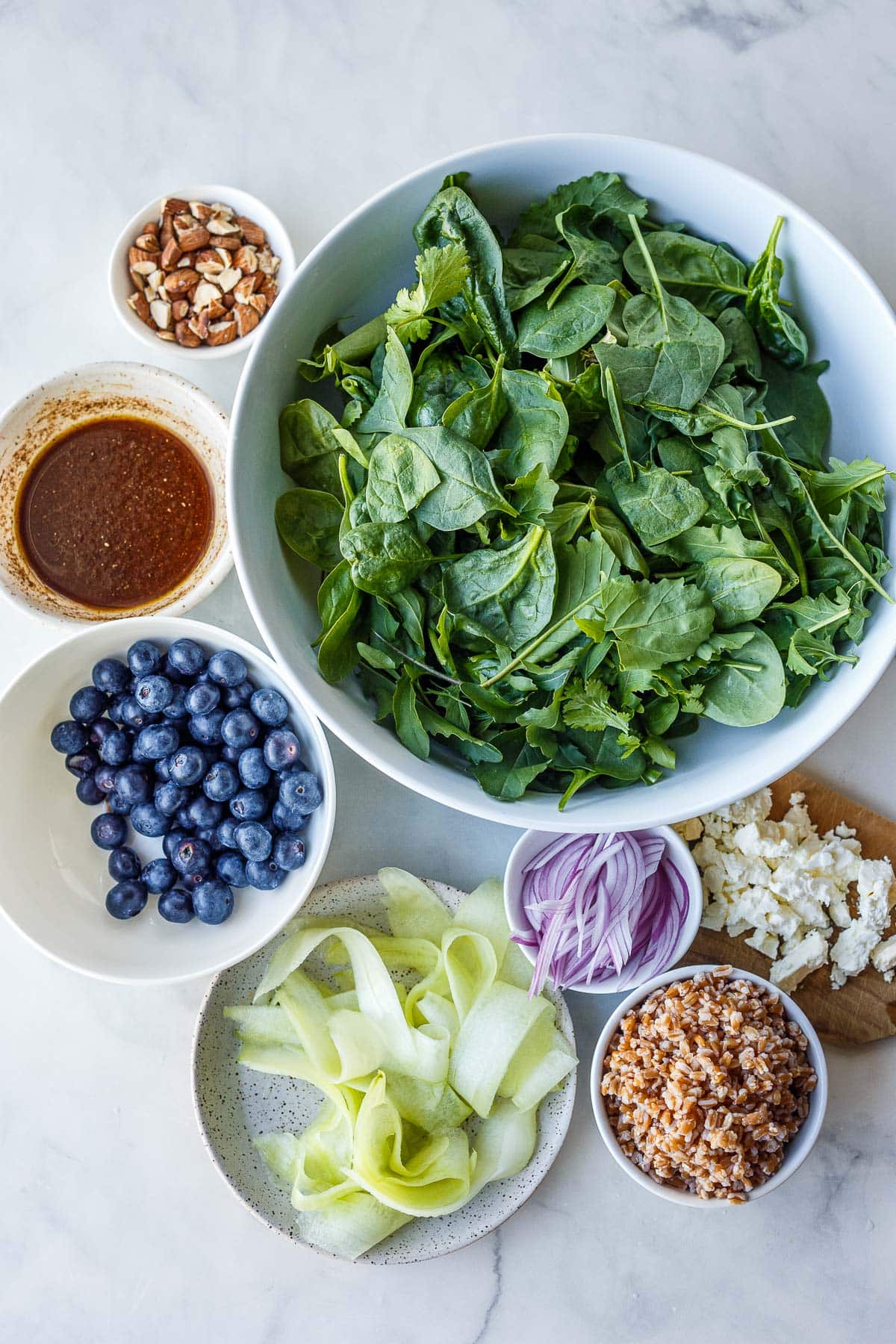ingredients for blueberry salad arranged neatly in bowls and ready to assemble - bowl of spinach, almonds, white balsamic dressing, blueberries, shaved cucumber, sliced red onion, farro, and crumbled feta.