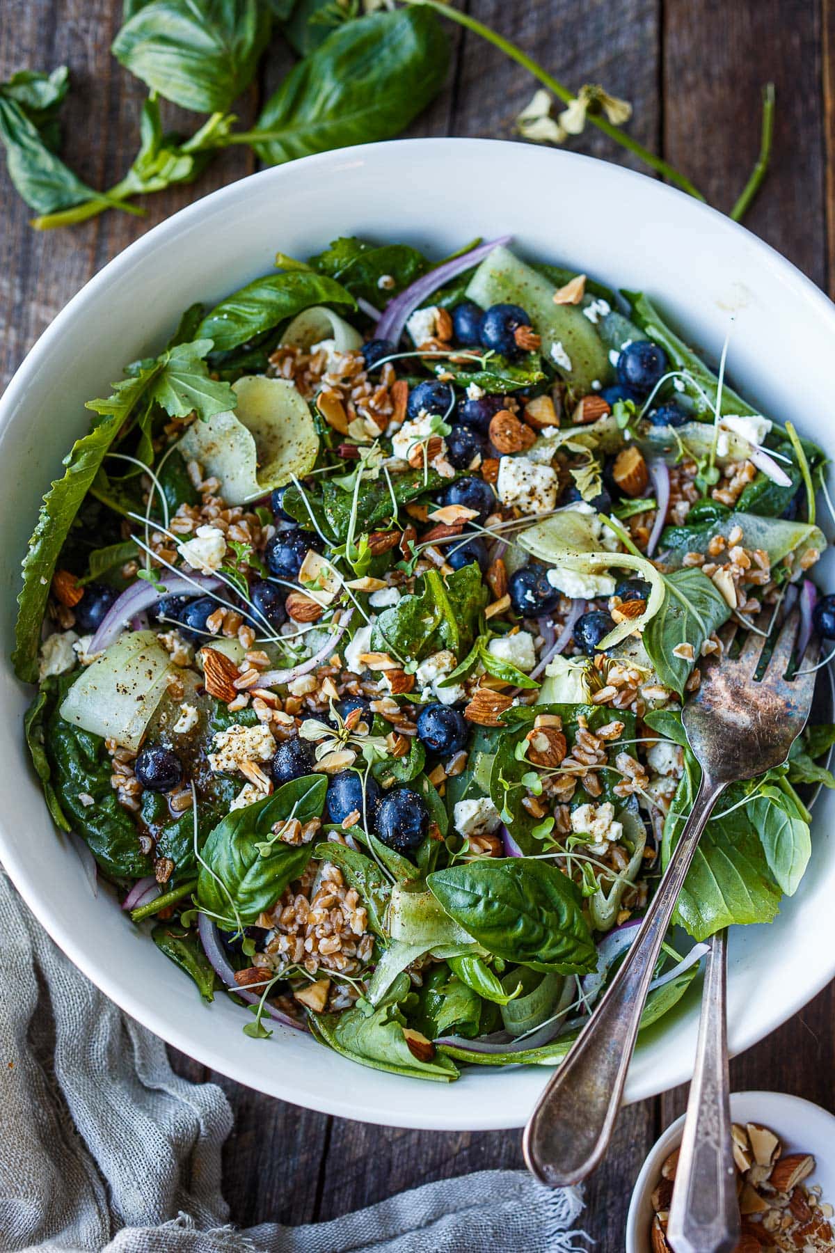 blueberry salad with summer greens, toasted almonds, shaved cucumber, sliced red onion, crumbled feta, and microgreens with a silver fork in the serving bowl.