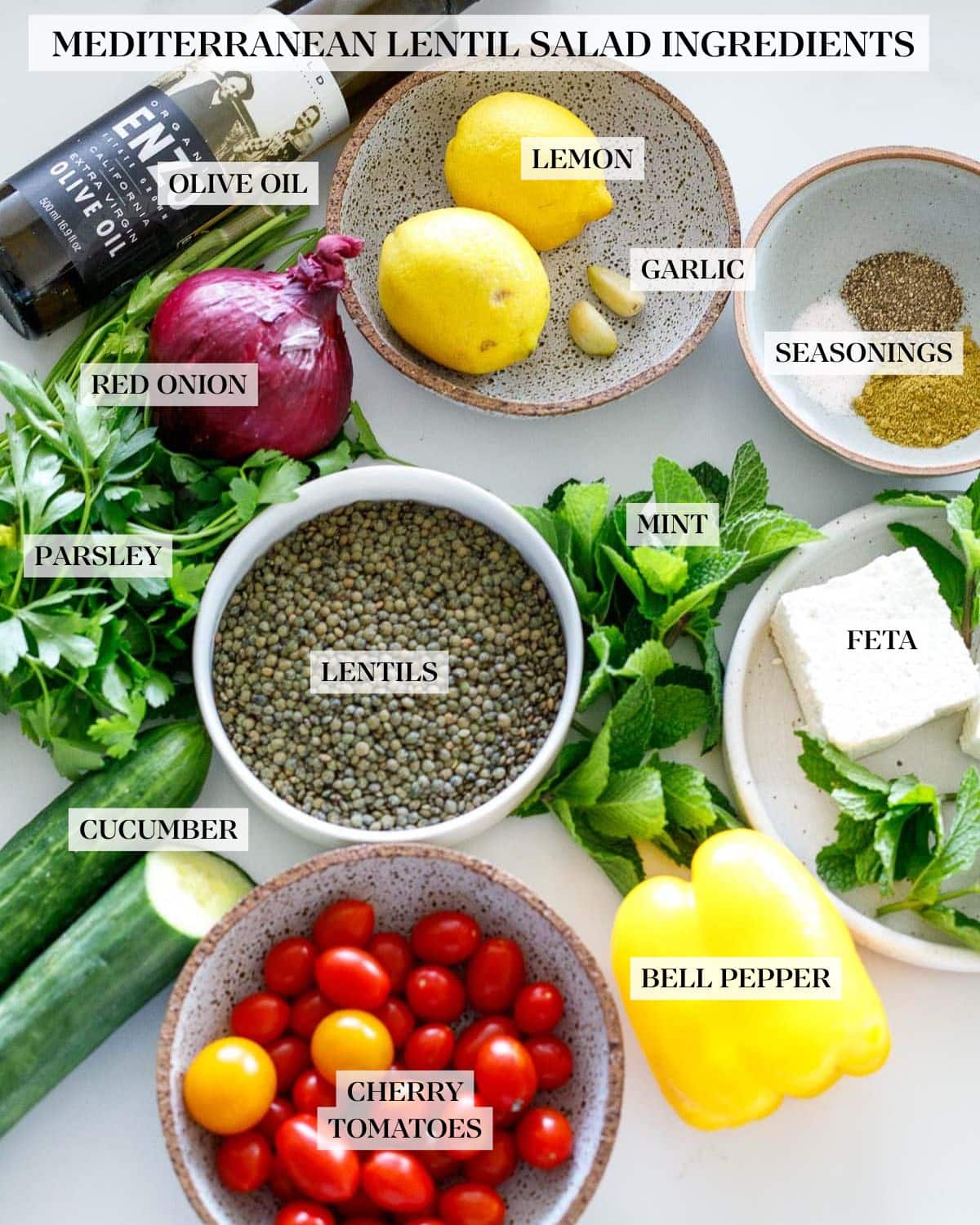 ingredients for lentil salad laid out in bowls - lemons, garlic cloves, spices, feta, herbs, lentils, cherry tomatoes, olive oil, red onion, cucumber.