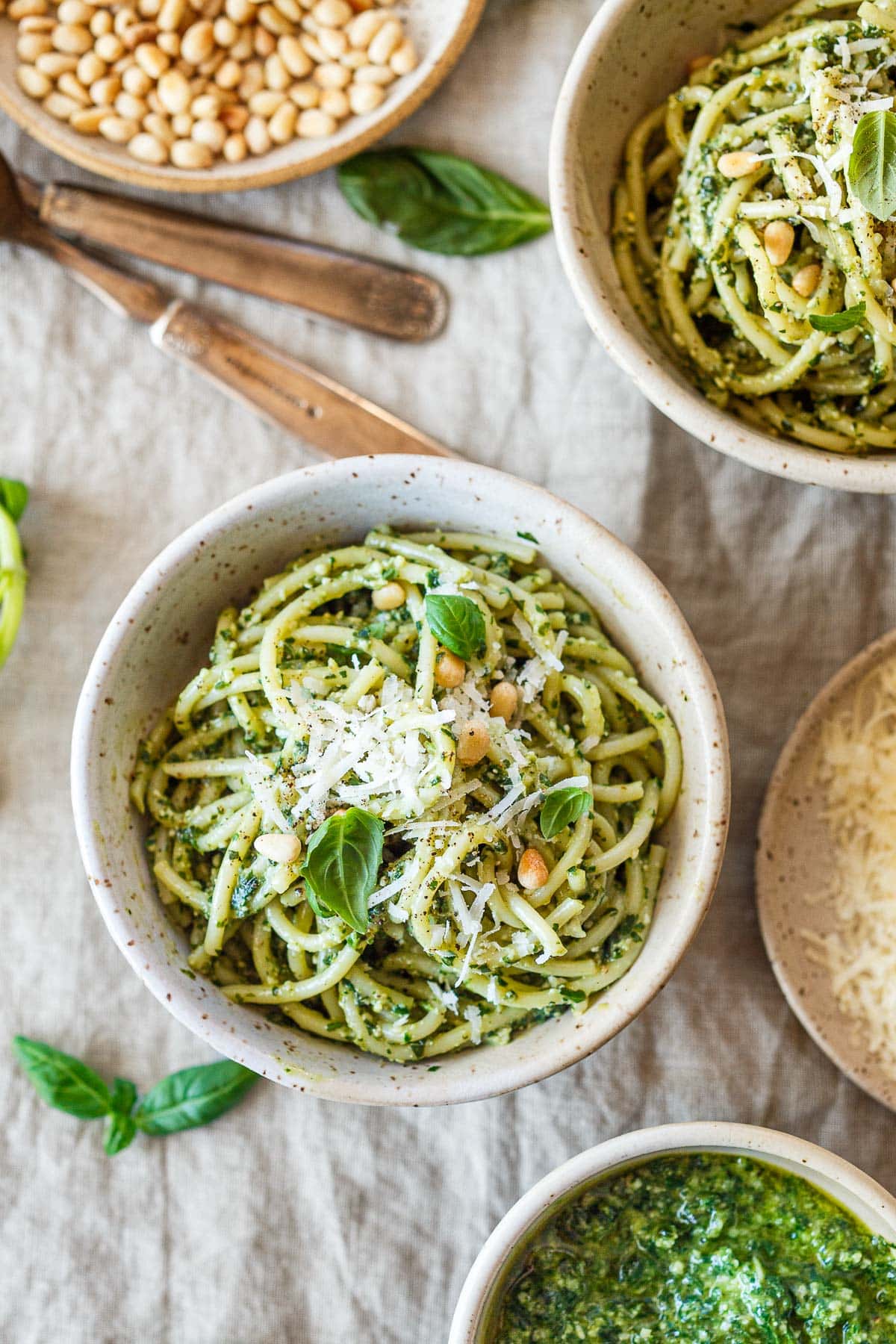 bowl of pesto pasta garnished with pine nuts, parmesan, and basil on linen cloth with bowls nearby with pine nuts, more pesto pasta, and pesto sauce.