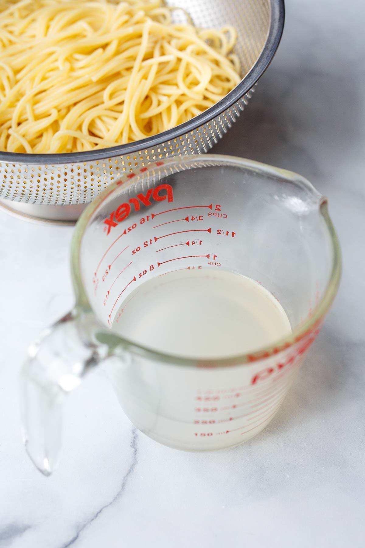 Metal colander with cooked spaghetti. A glass pyrex cup of pasta water.