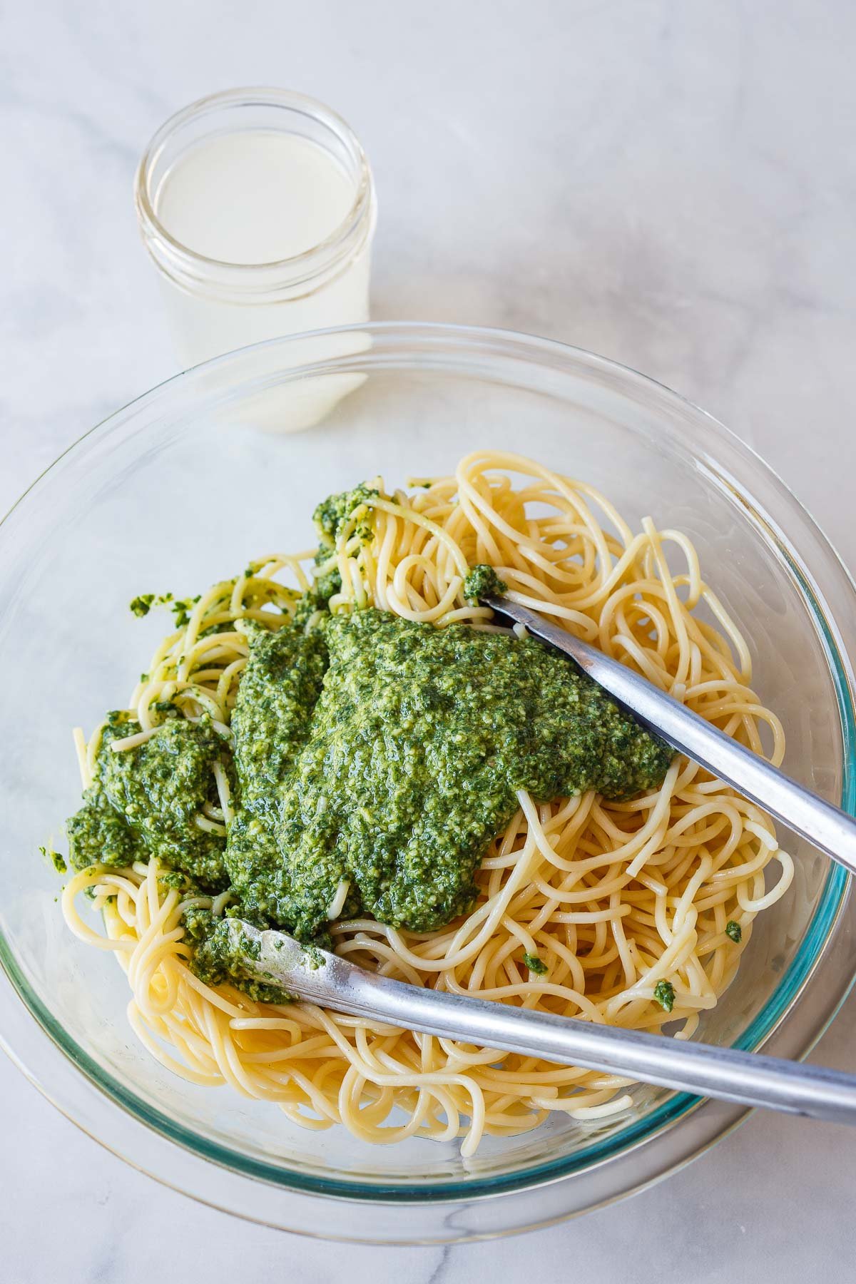 glass mixing bowl with spaghetti and pesto sauce over top with tongs to mix next to jar of pasta water.