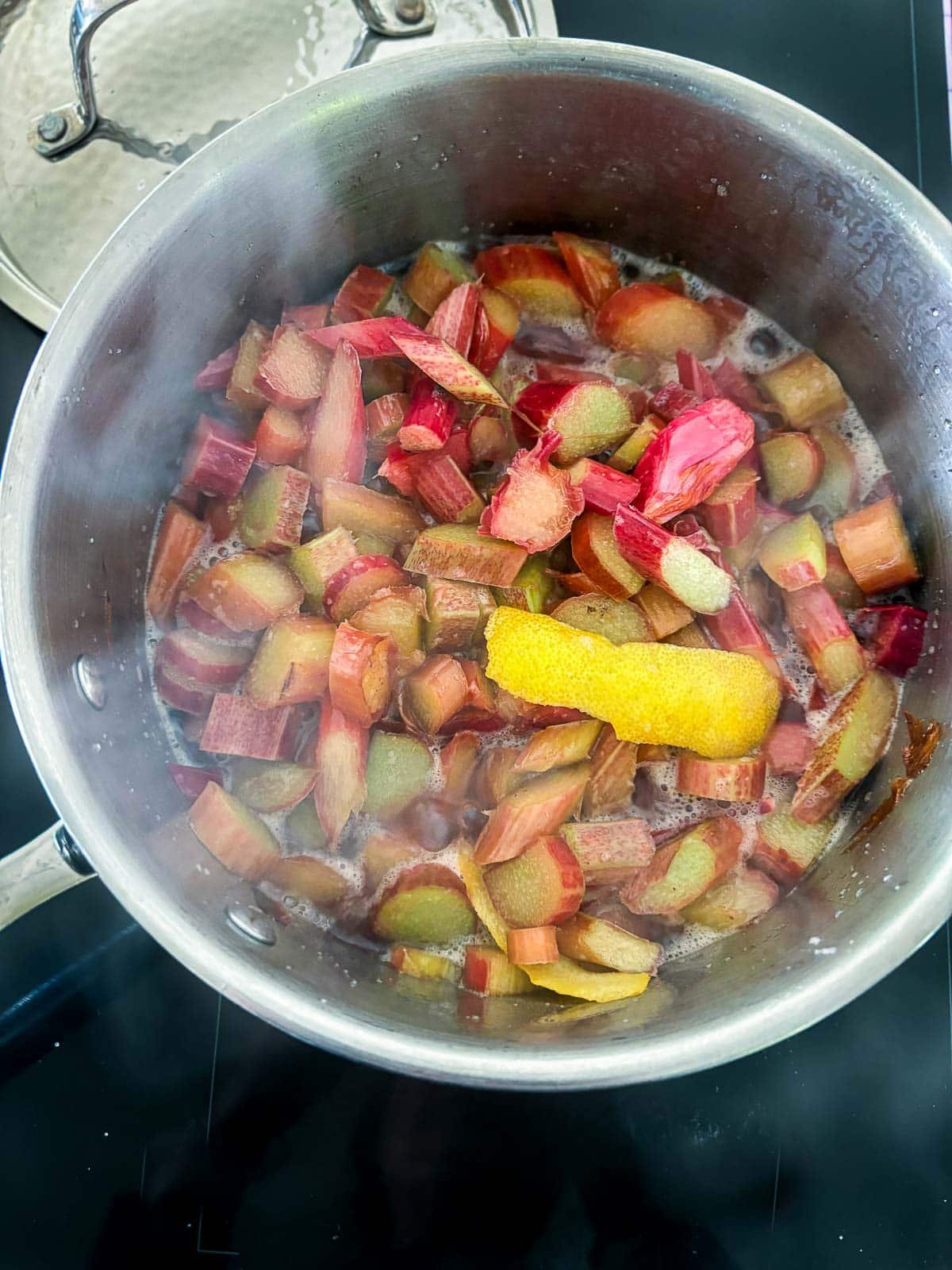 pot on stovetop with ingredients to make rhubarb compote simmering, including rhubarb pieces and lemon peels.