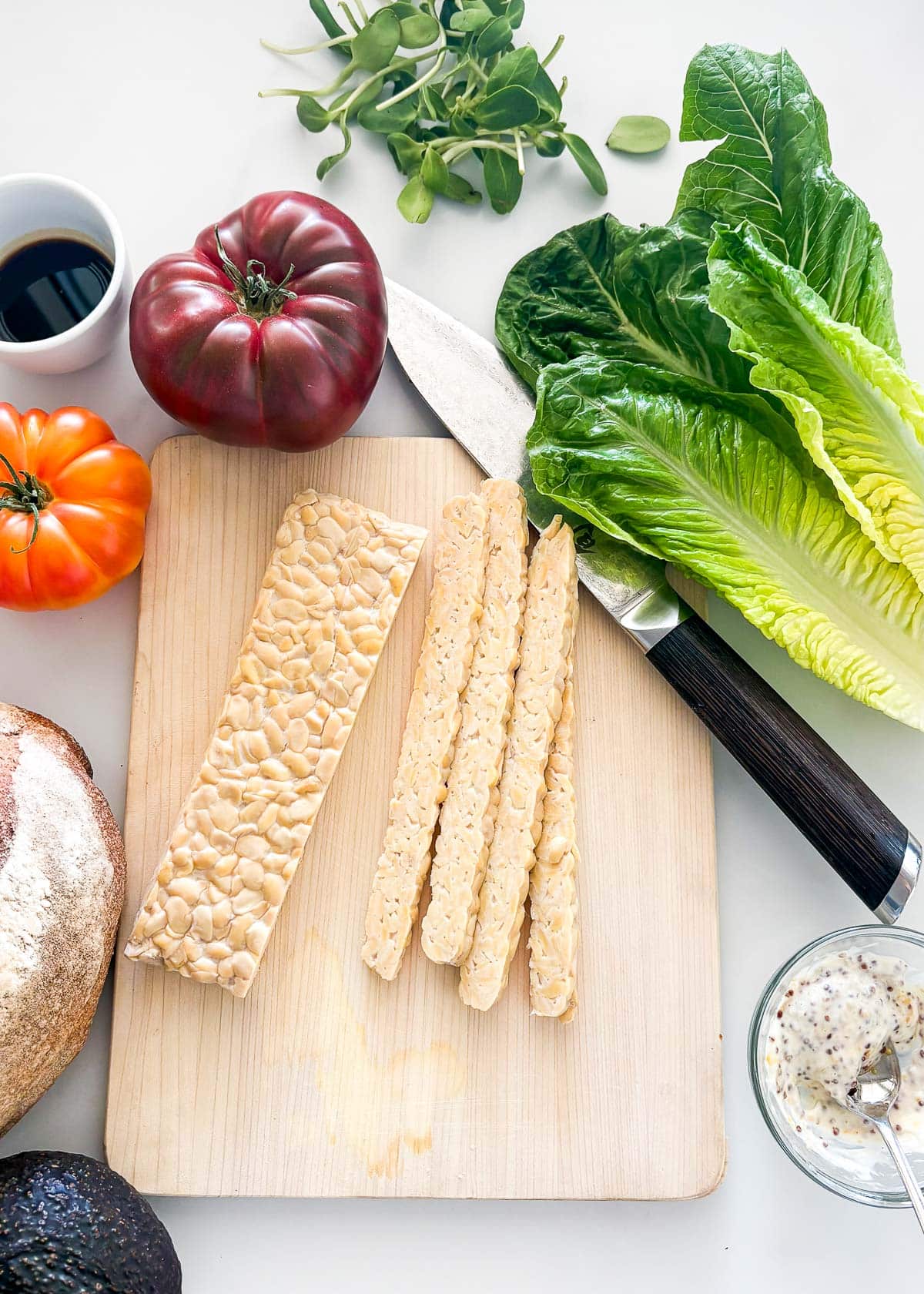 ingredients for BLT sandwich - sprouts, romaine, bowl of soy sauce, heirloom tomatoes, cutting board and knife with sliced tempeh, sourdough, avocado, bowl of mayo and whole grain mustard. 