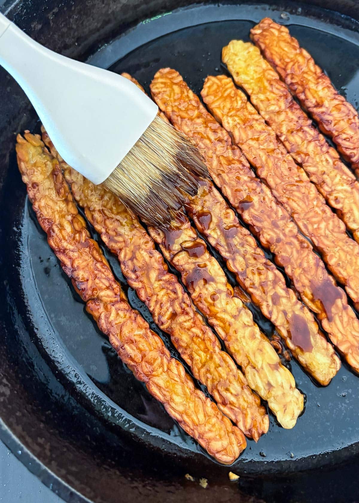 kitchen brush brushing maple soy glaze over tempeh bacon in cast iron skillet. 