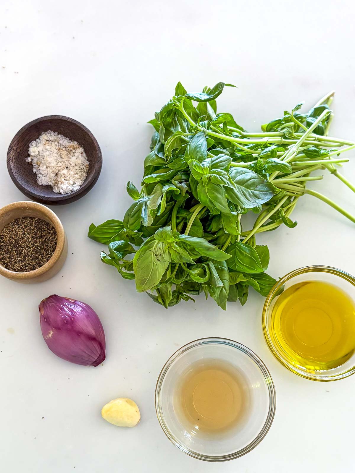 bunch of fresh basil, shallot, garlic clove, and small bowls with salt, pepper, vinegar, and olive oil, arranged neatly to make basil vinaigrette.