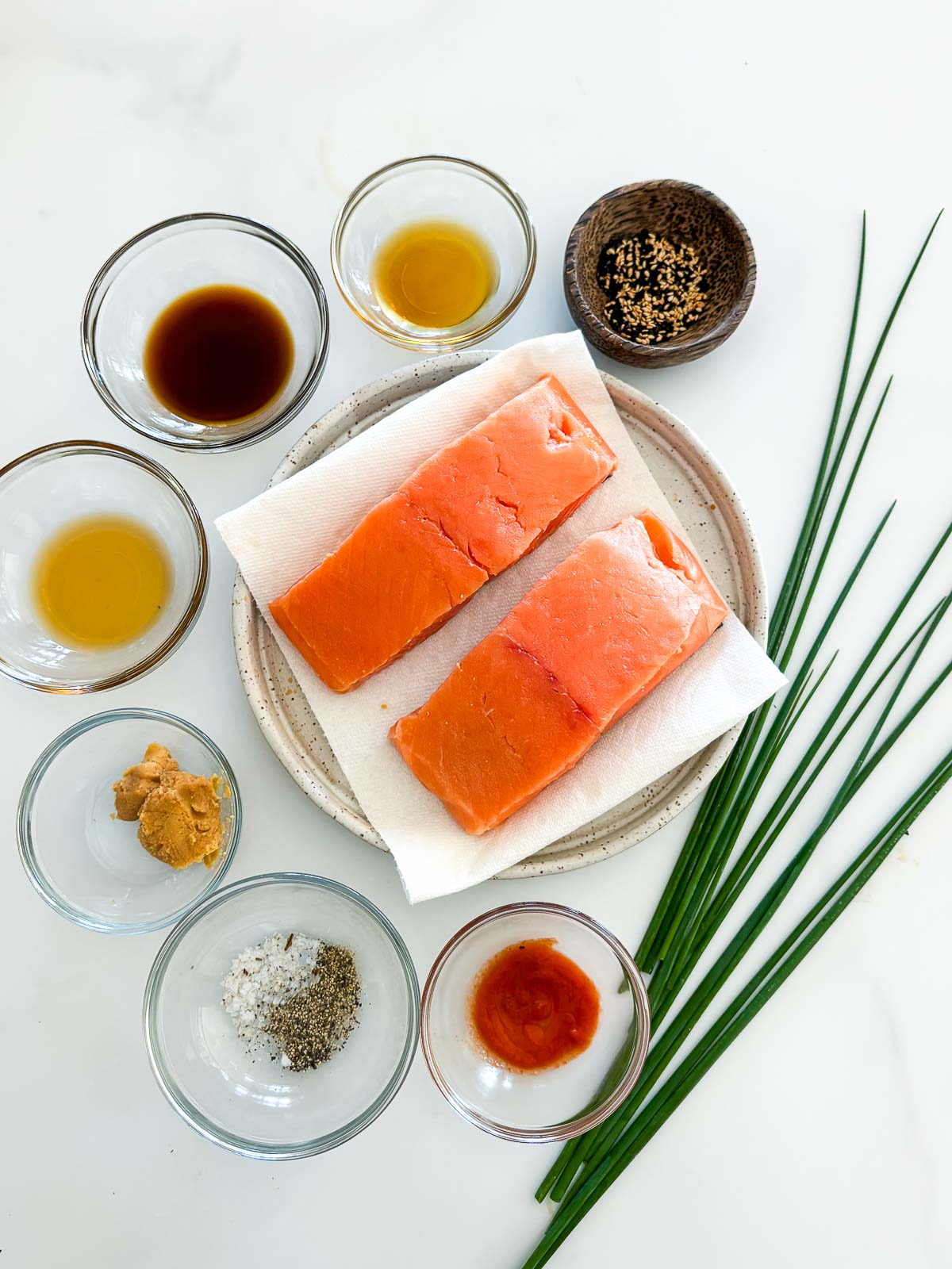 ingredients laid out for broiled salmon - two salmon filets on a plate, surrounded by small bowls of sesame seeds, sesame oil, soy sauce, olive oil, miso paste, salt and pepper, and sriracha, next to chives.