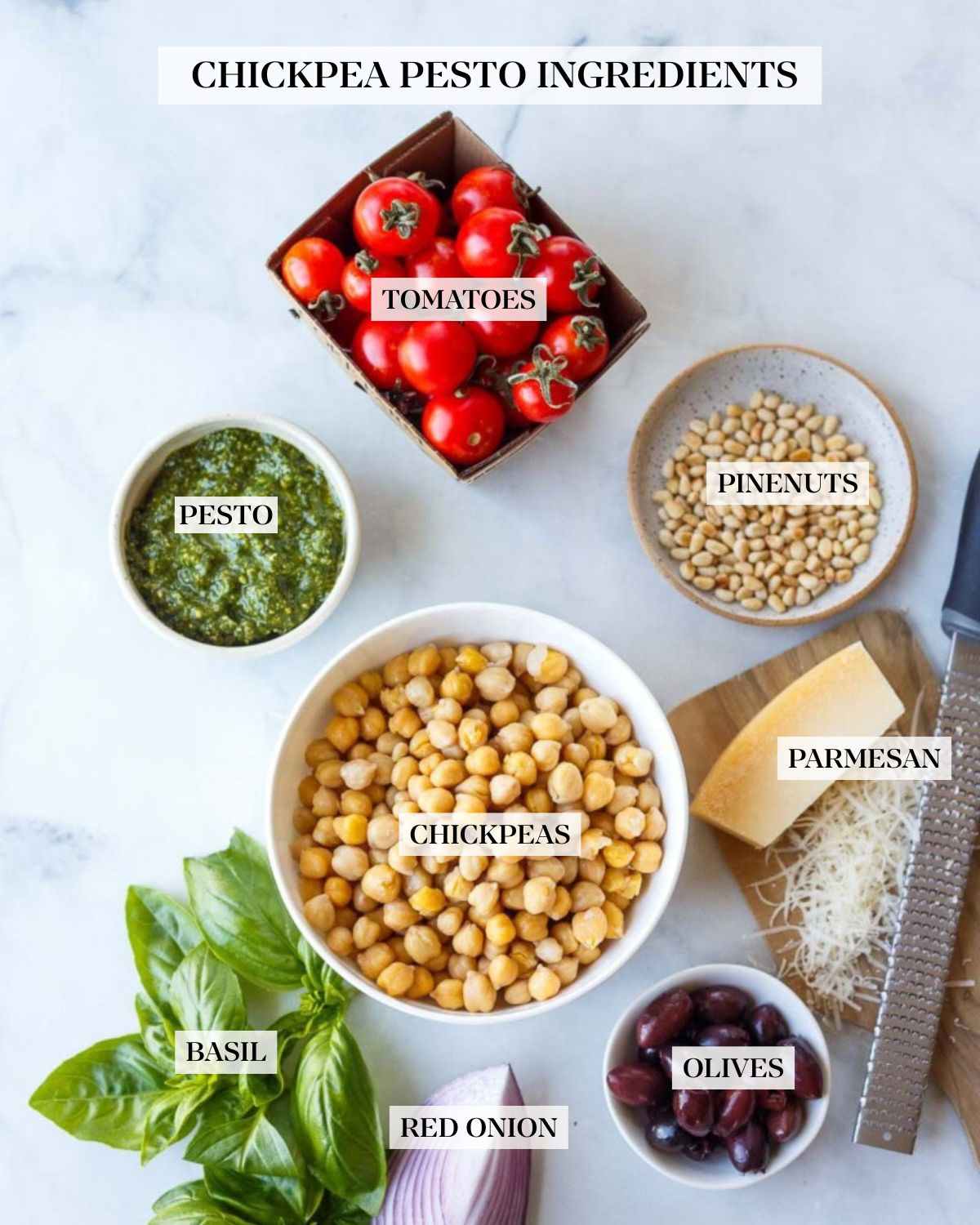 ingredients arranged for making pesto chickpeas - carton of cherry tomatoes, small bowls of pine nuts and pesto, bowl of chickpeas, grating parmesan on small wood board, small bowl of olives, wedge of red onion, and fresh basil bunch.