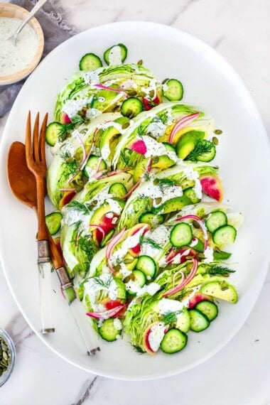 oval platter with wedge salad with red onion, cucumber, avocado, pepitas, watermelon radish, and fresh dill, topped with creamy dressing, served with wood utensils.