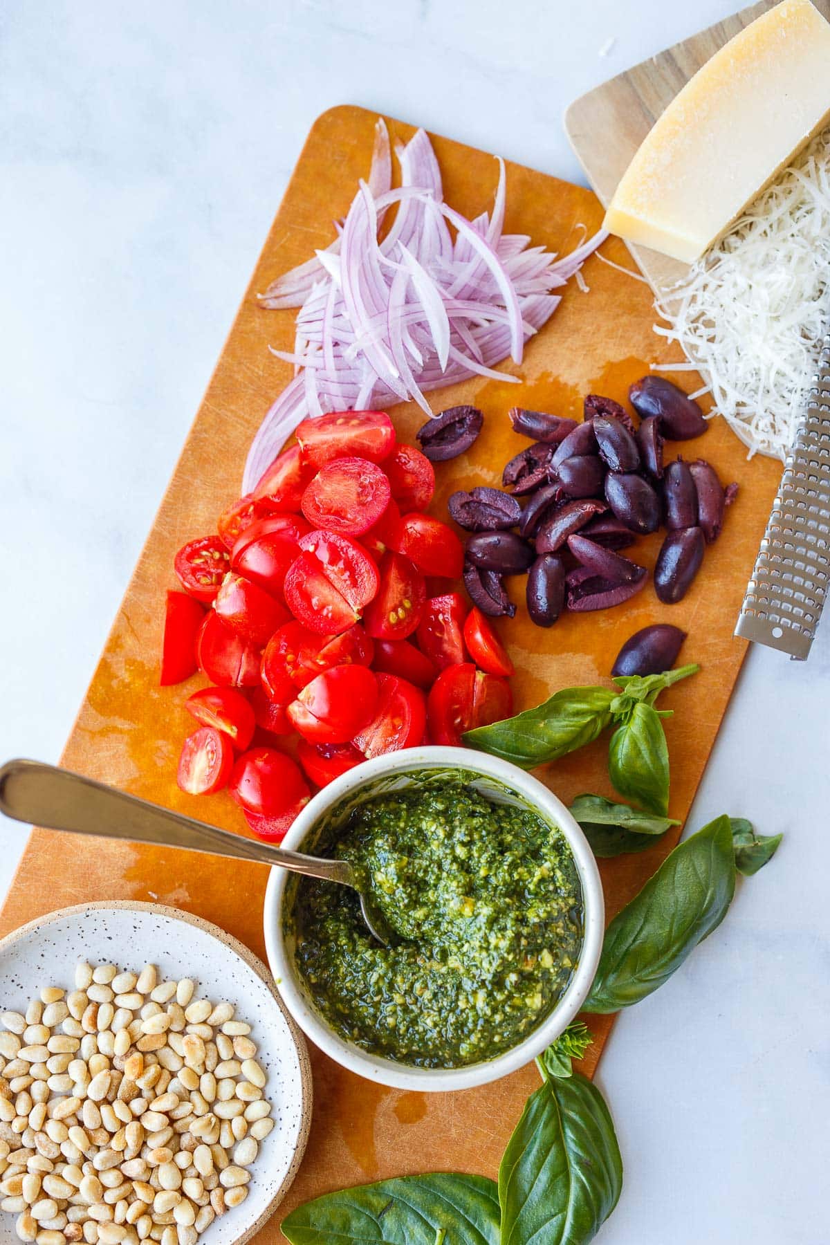 narrow cutting board with pesto chickpeas ingredients prepped - thin red onion slices, grated parmesan, quartered olives and cherry tomatoes, bowl of pesto and pine nuts, and fresh basil leaves.