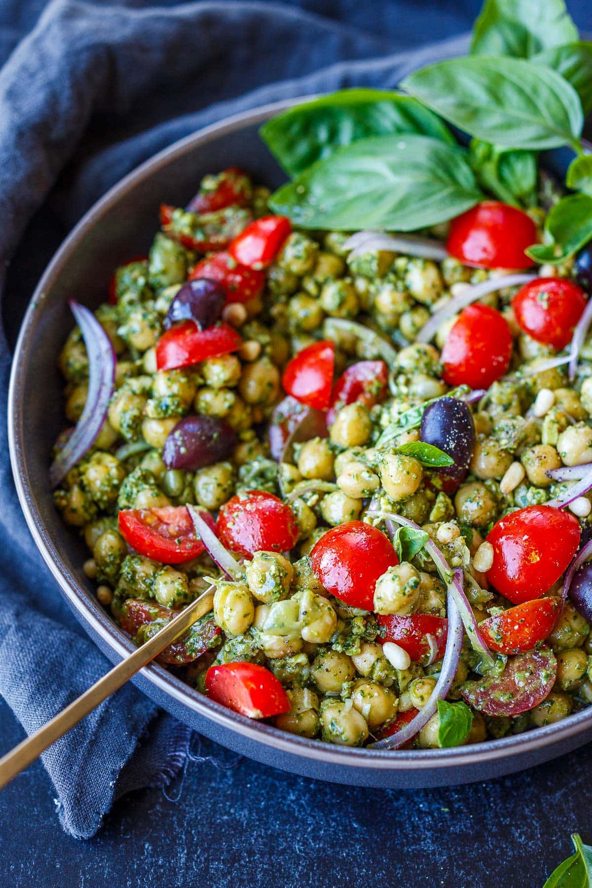 pesto chickpeas in bowl with pine nuts, kalamata olives, cherry tomatoes, thin red onion slices, and basil.