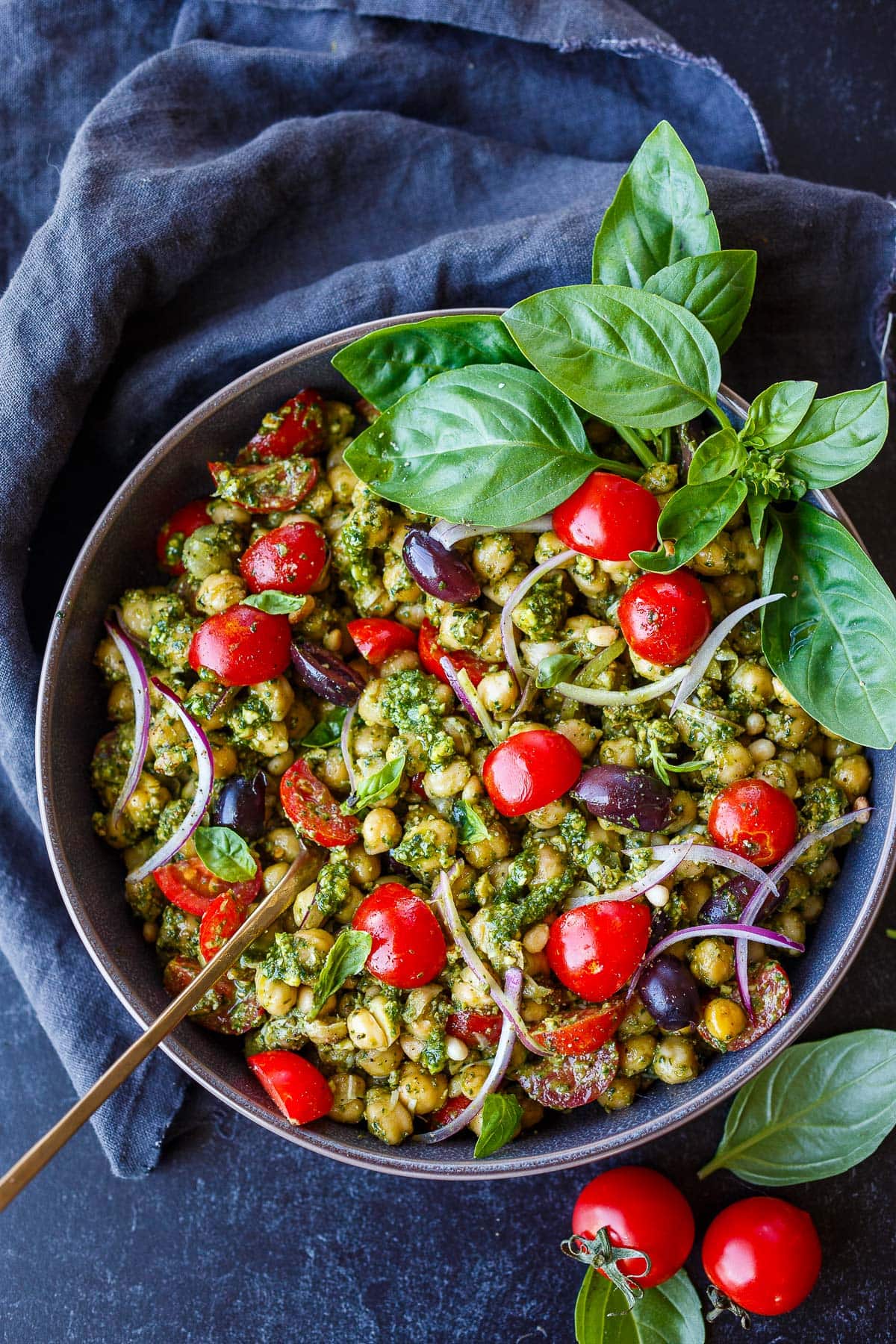 bowl of pesto chickpeas topped with cherry tomatoes, red onion slices, kalamata olives, and fresh basil leaves.