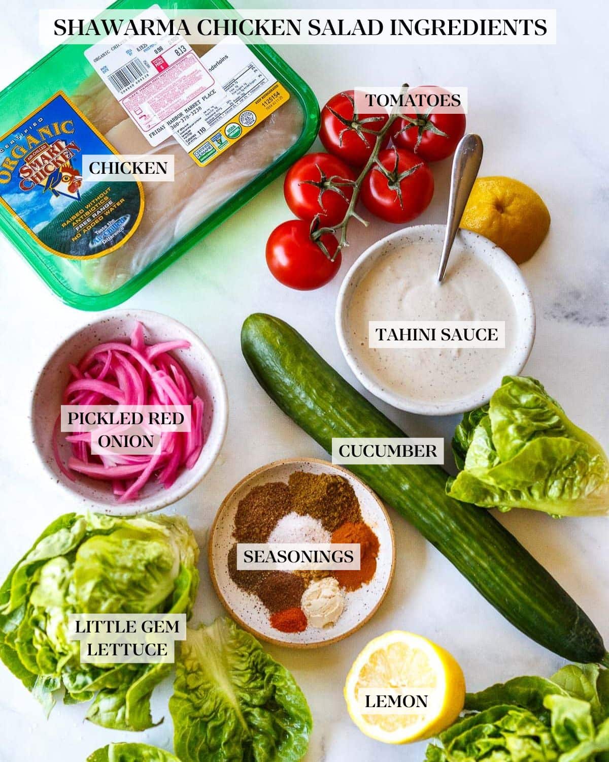 chicken shawarma salad ingredients arranged neatly on counter - packaged chicken, tomato on the vine, lemon half, bowl of tahini yogurt dressing, little gem lettuce, cucumber, bowl of pickled onions, small dish of shawarma spices. 