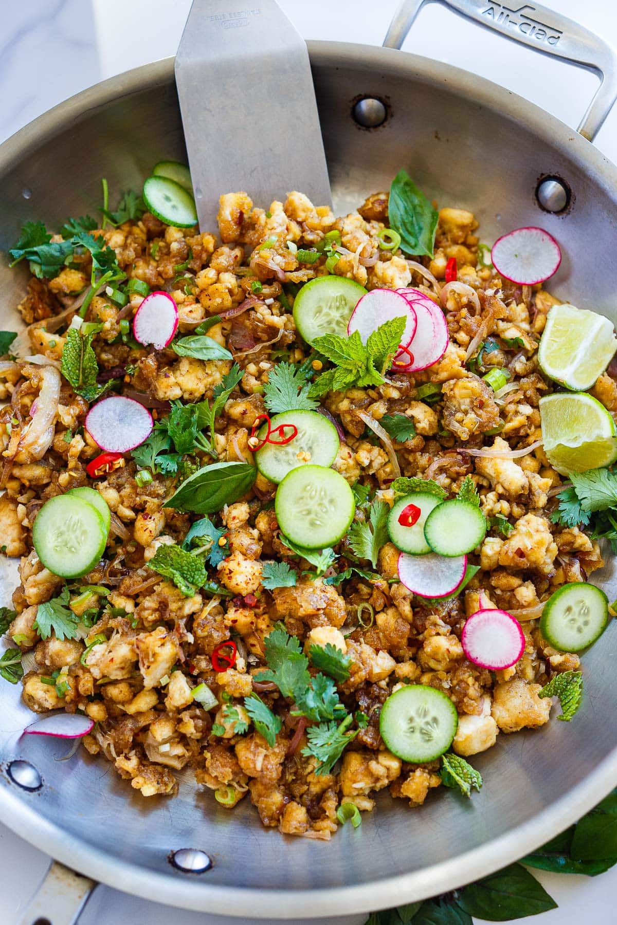Thai tofu larb in skillet garnished with sliced cucumber, radish, and chilies, fresh herbs, and lime wedges. 