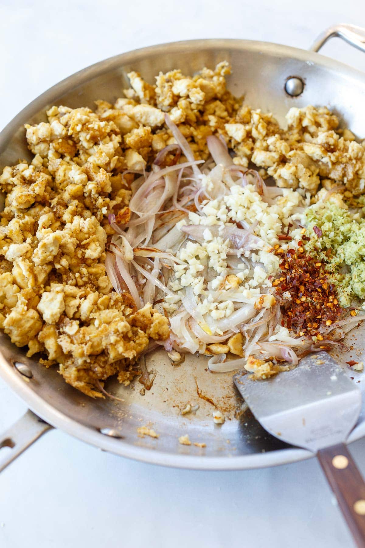 skillet with aromatics in center- garlic, ginger, shallot, chili flakes, and lemongrass, surrounded by tofu larb, with metal spatula. 