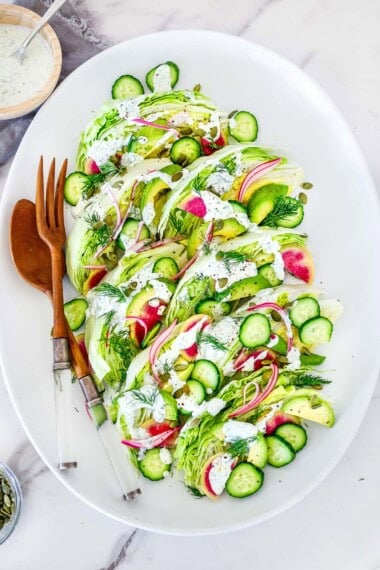 oval platter with wedge salad with red onion, cucumber, avocado, pepitas, watermelon radish, and fresh dill, topped with creamy dressing, served with wood utensils.