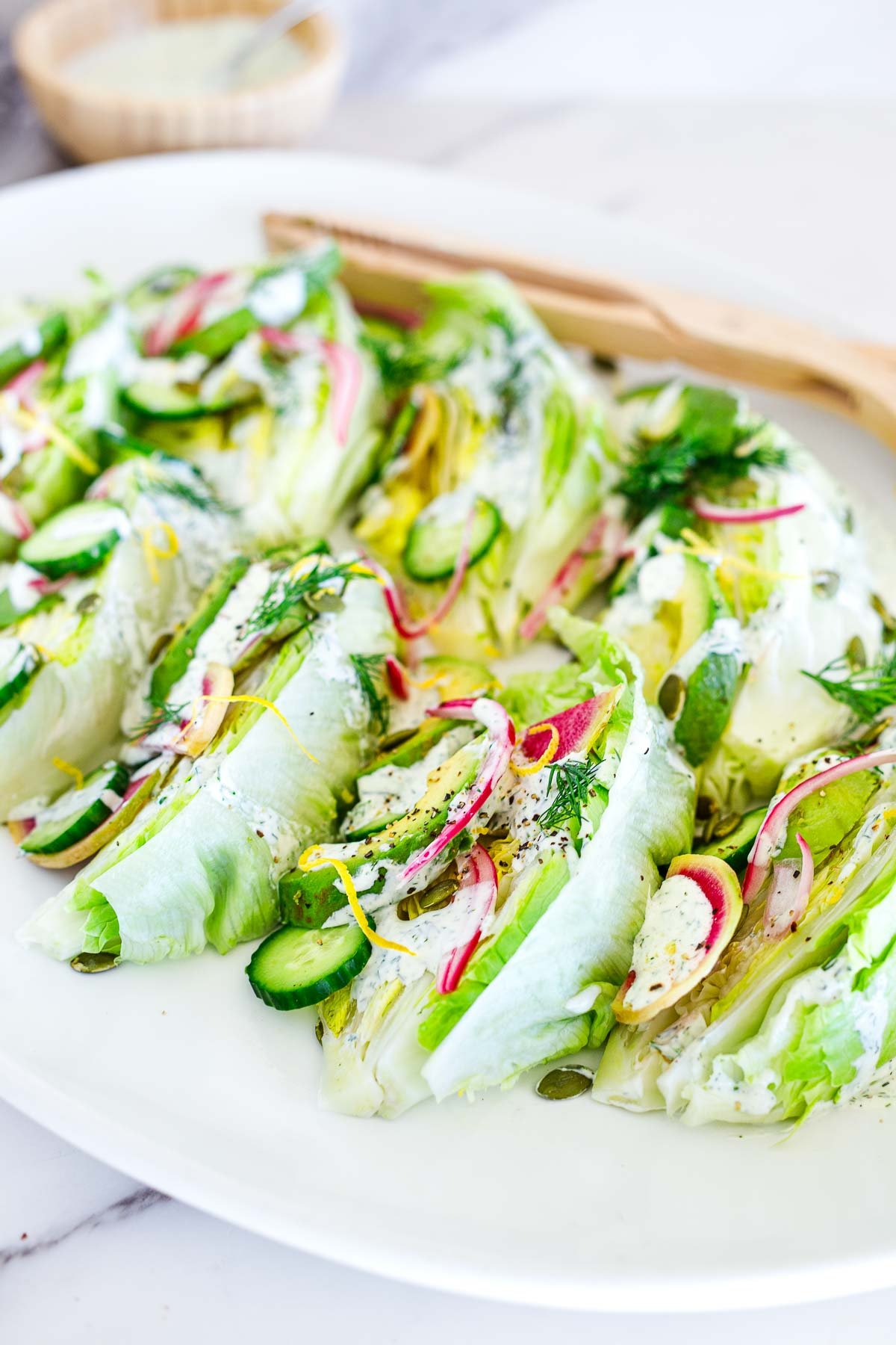 serving platter with wood utensils and iceberg lettuce salad topped with dilly ranch dressing, fresh dill, cucumber, radish, onion, pepitas, avocado. 