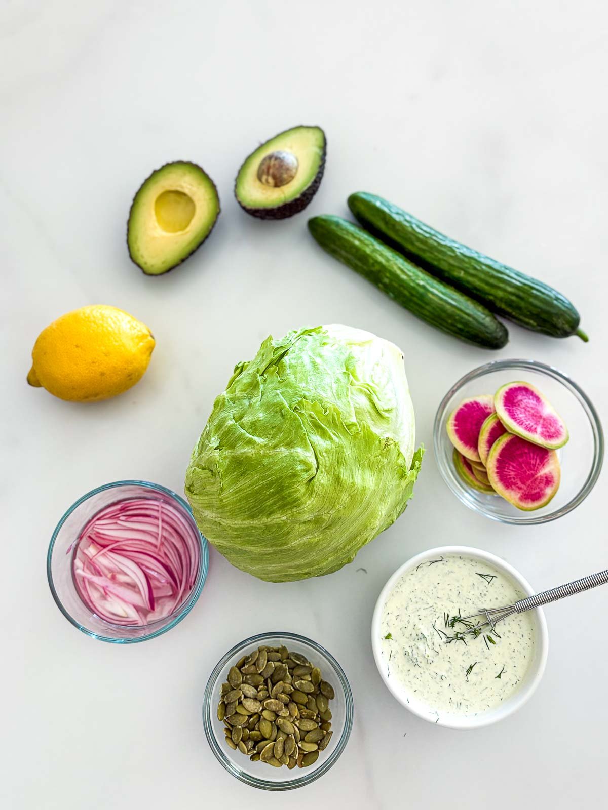 ingredients laid out for wedge salad - avocado, cucumber, watermelon radish, dilly ranch dressing, iceberg lettuce, pepitas, red onion, and lemon. 