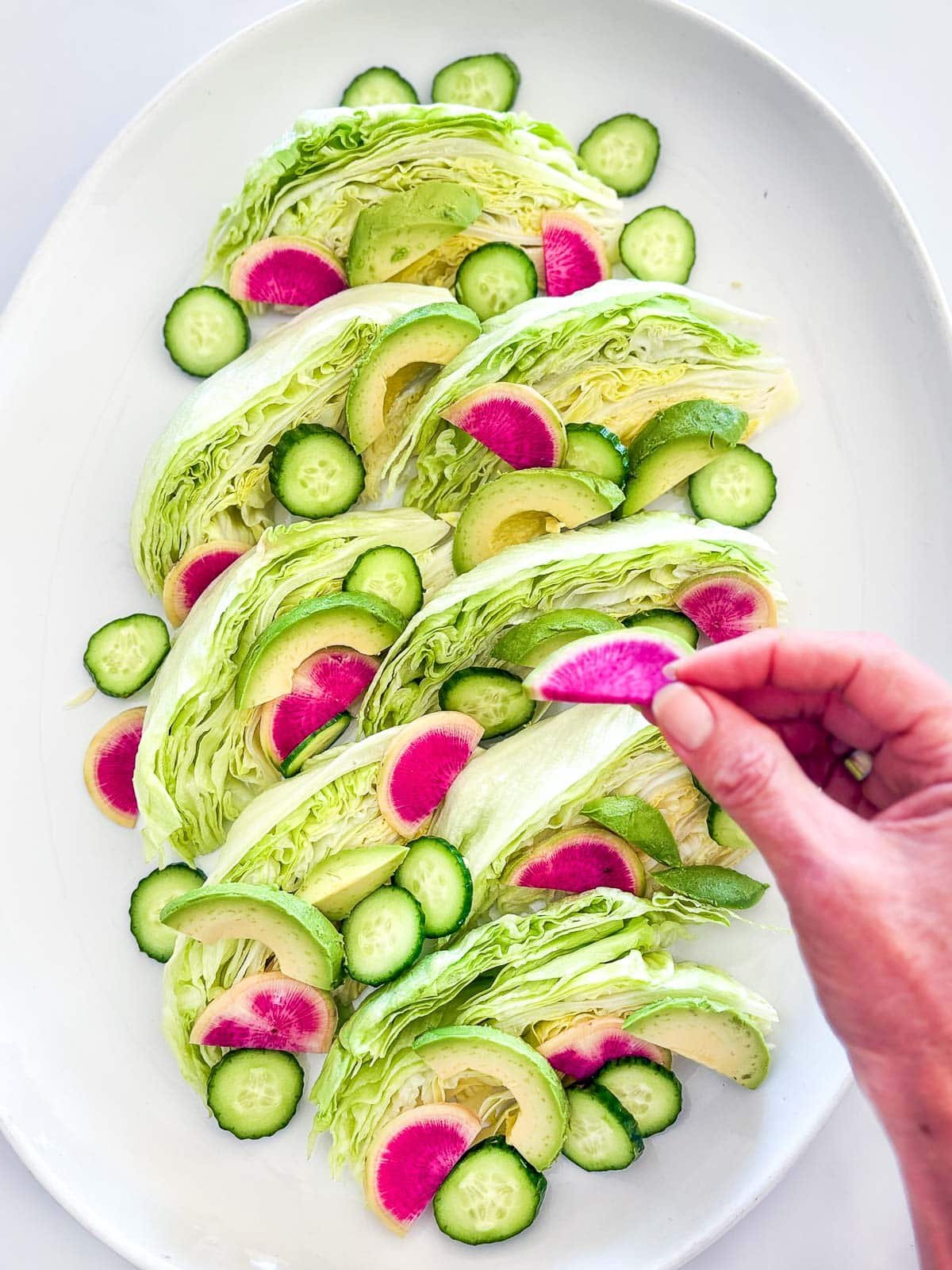 hand placing watermelon radish on wedges of iceberg lettuce, with cucumbers and avocados also placed on the salad. 