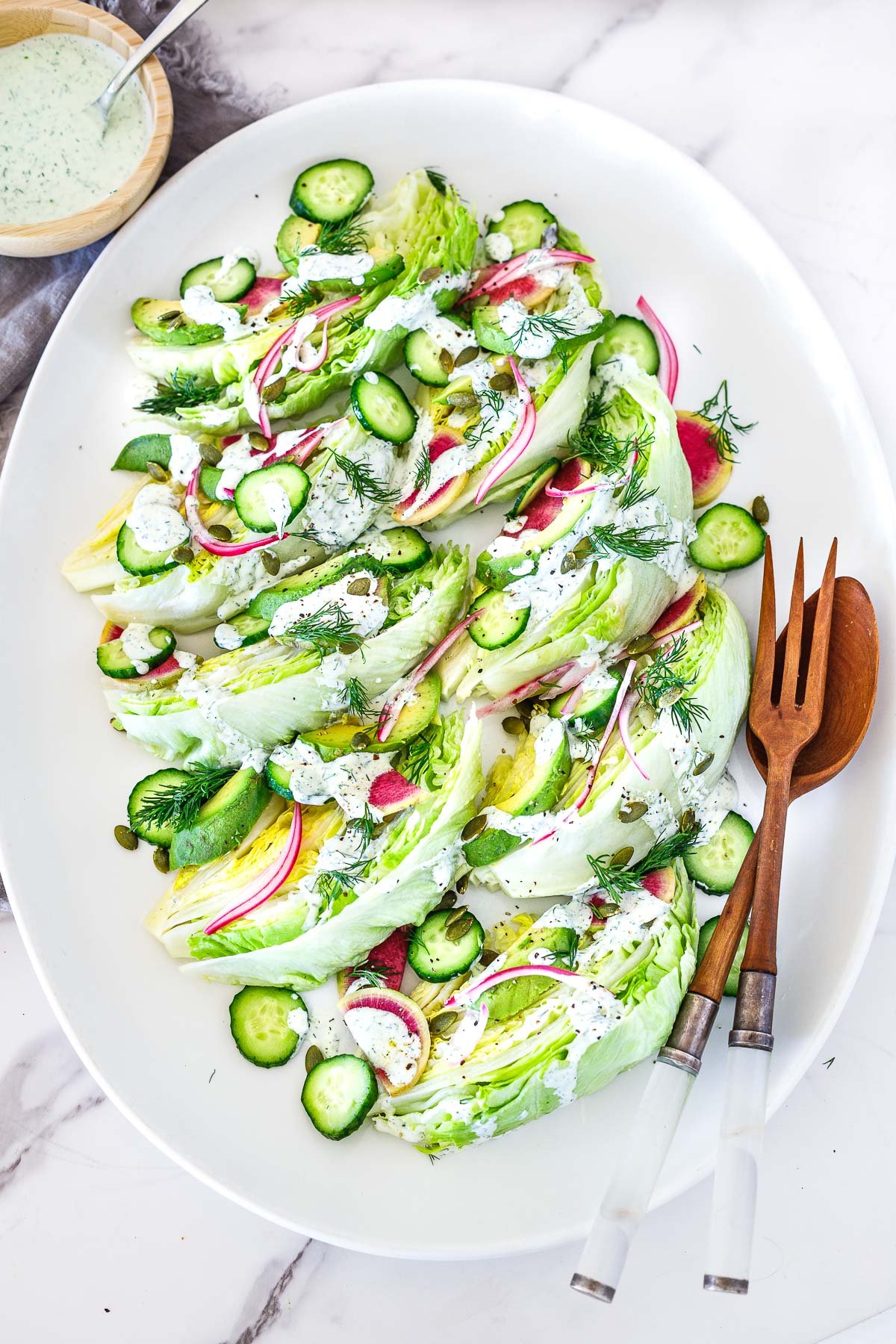 serving platter with wood utensils and an iceberg lettuce salad topped with creamy ranch dressing, fresh dill, cucumber, radish, onion, pepitas, avocado. 