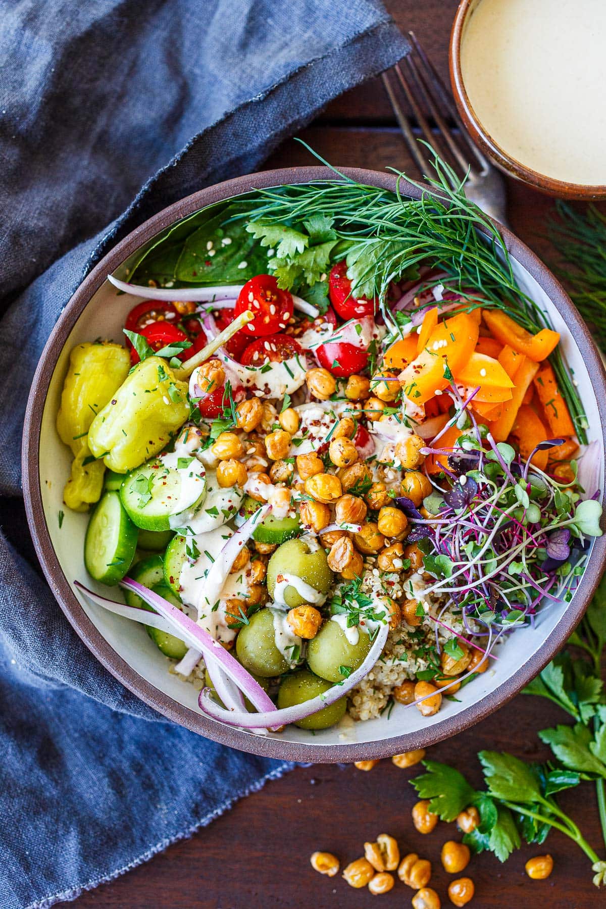 Mediterranean chickpea bowl with crispy chickpeas in the center, colorful veggies around, tomatoes, peppers, micro greens, olives, red onion, cucumber, pepperoncini, and herbs, topped with tahini sauce.