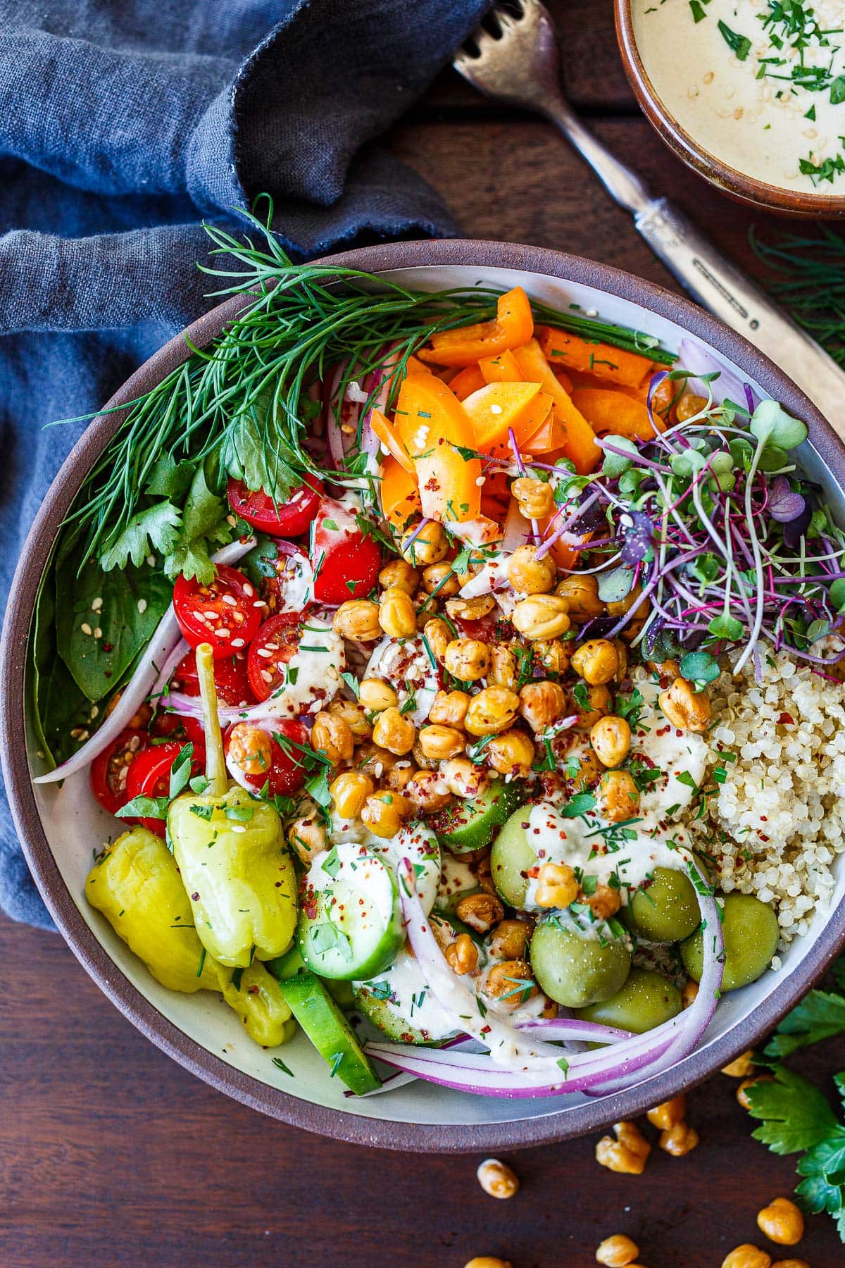 Crispy chickpea bowl with quinoa, topped with micro greens, orange bell pepper, dill, parsley, basil, cherry tomatoes, red onion, pepperoncini, cucumber, olives, and chickpeas, with tahini sauce.