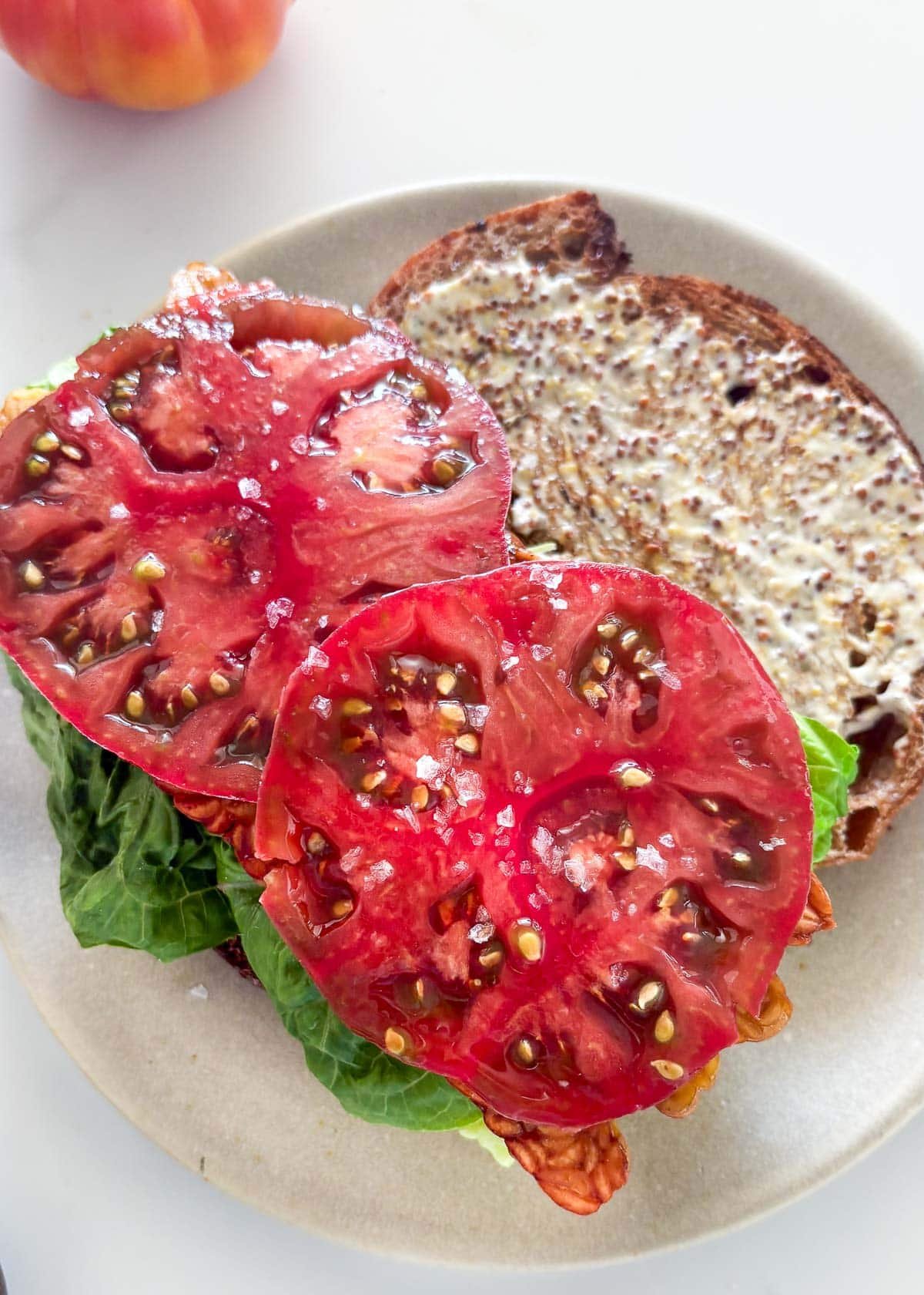assembling the BLT sandwich with mayo and mustard on one slice and lettuce and salted tomato on the other.