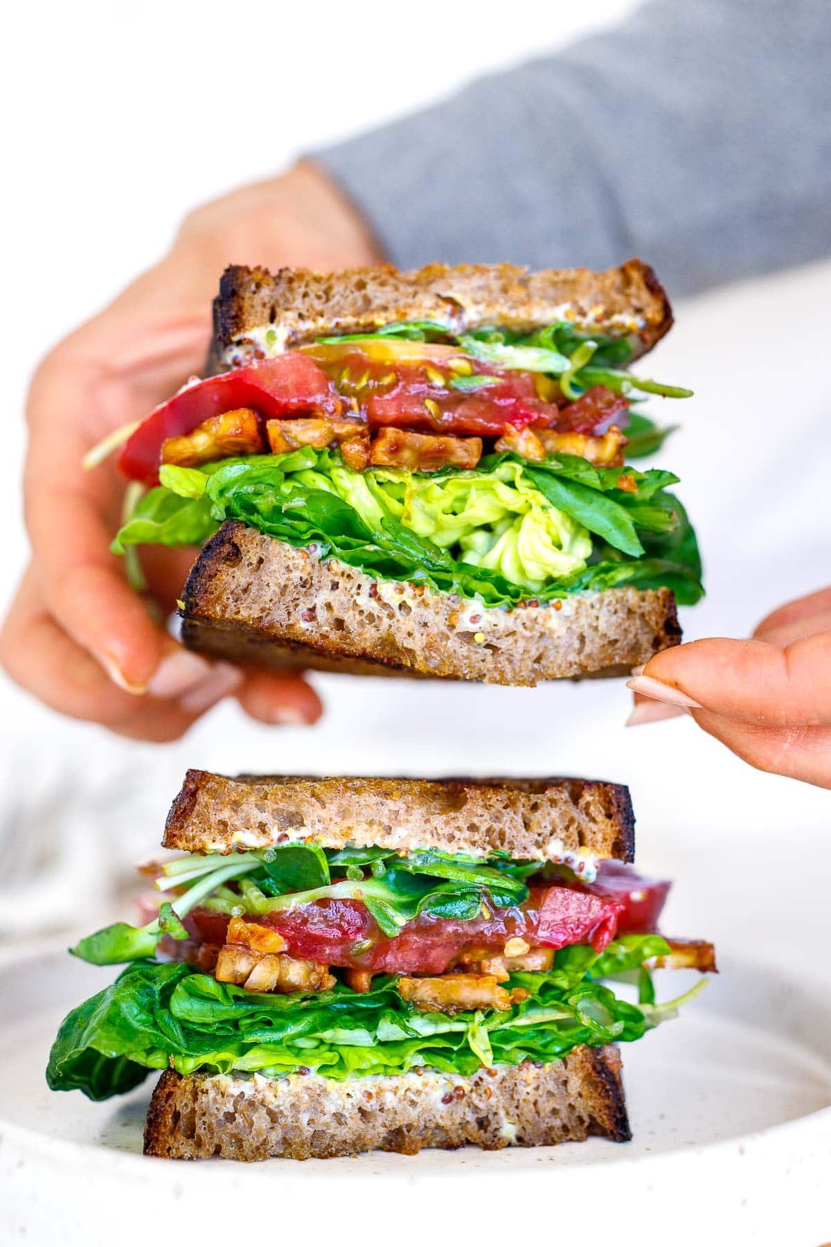 two BLT sandwiches with hands placing one sandwich to stack on top of the other, full of crisp lettuce, tempeh bacon, and tomato. 