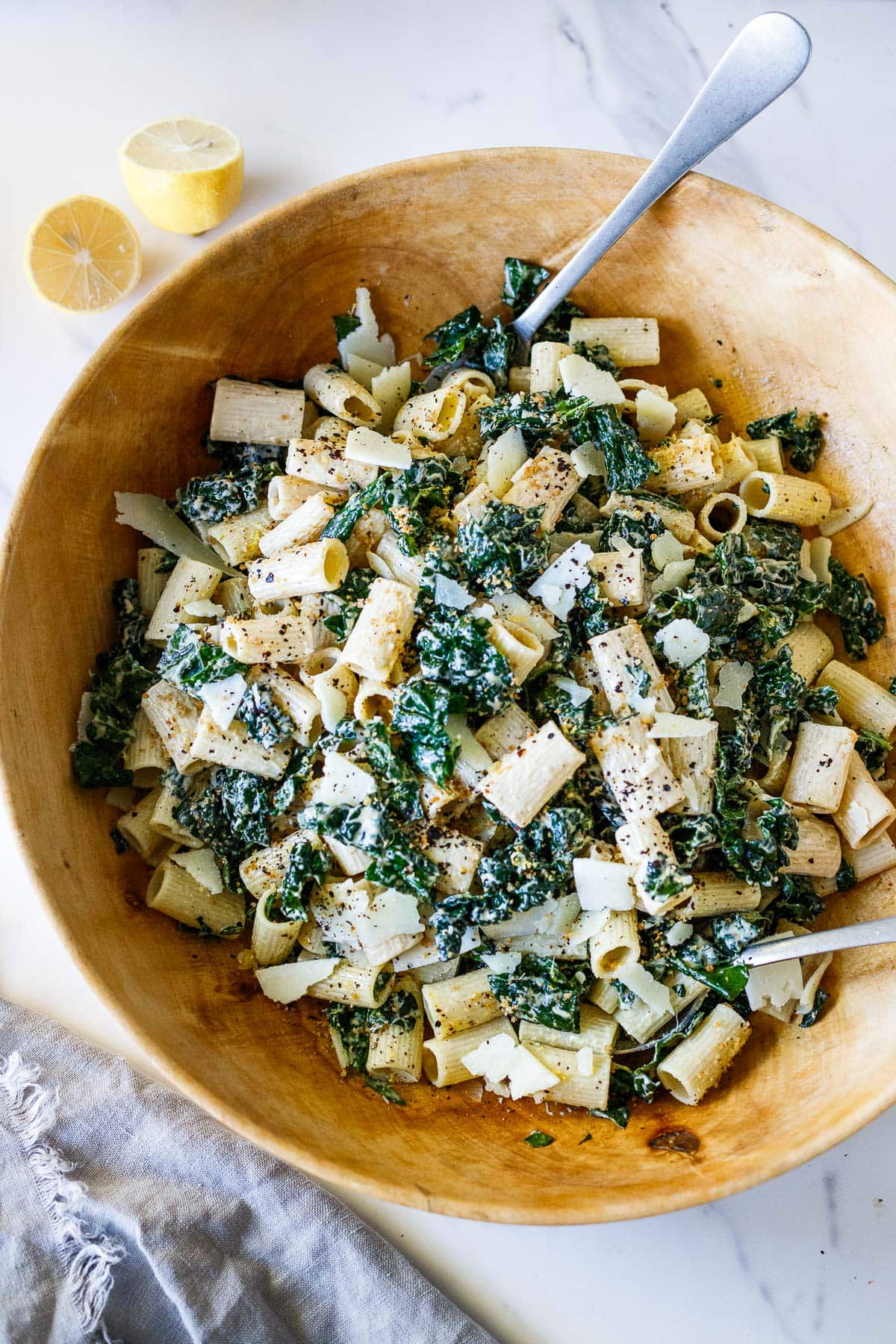 large wood bowl with kale caesar pasta salad with two silver utensils, garnished with pepper and chili flakes. 