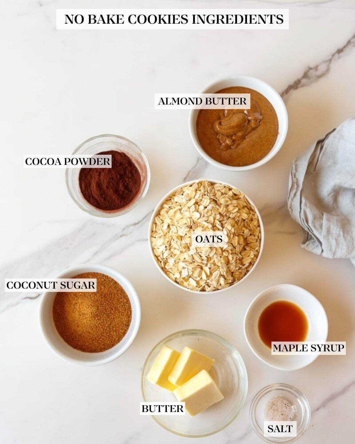 no bake cookies ingredients labeled and neatly arranged in bowls - almond butter, cocoa powder, oats, coconut sugar, butter, maple syrup, and salt. 