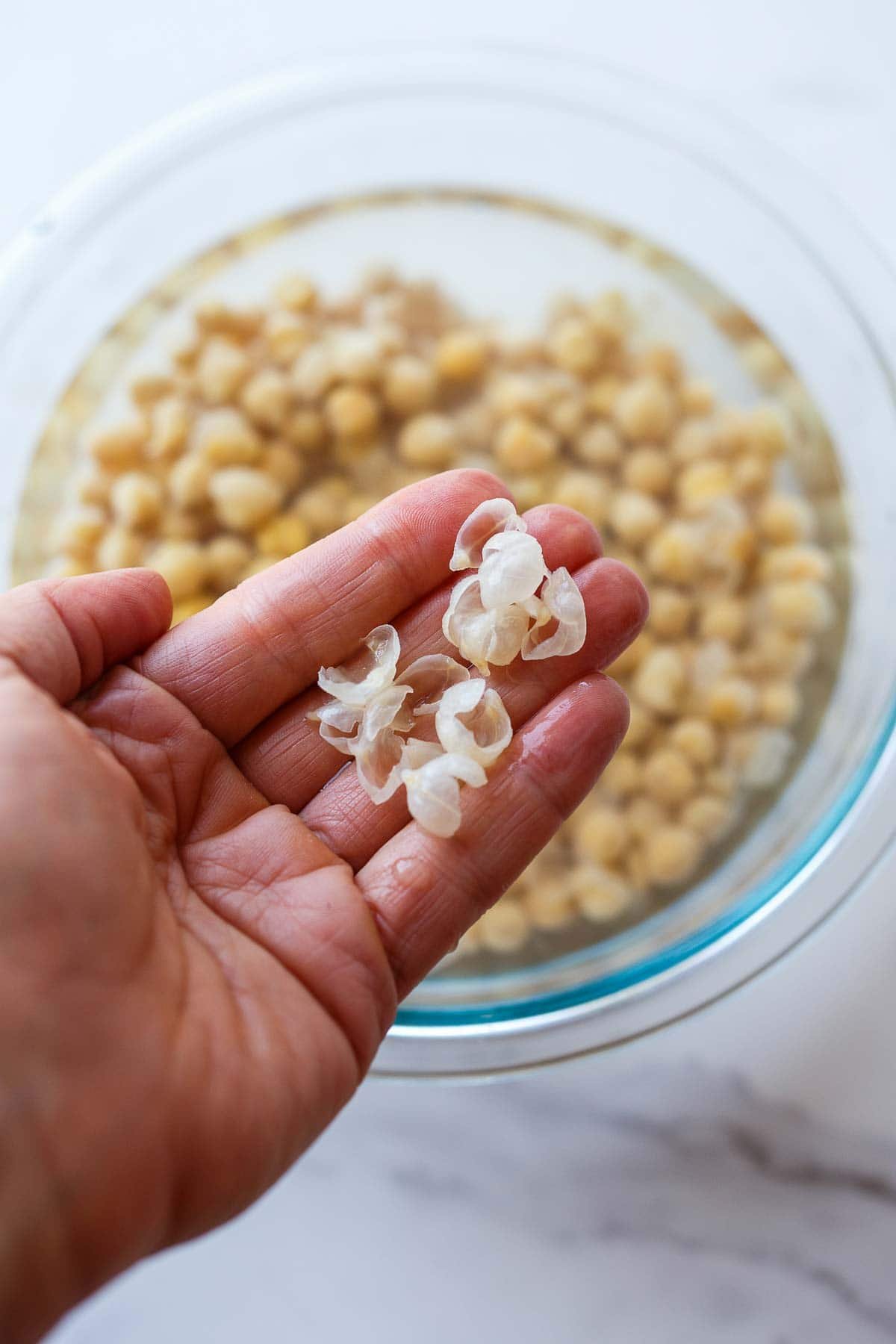 hand holding up peels that were removed from chickpeas, over bowl of chickpeas in water.