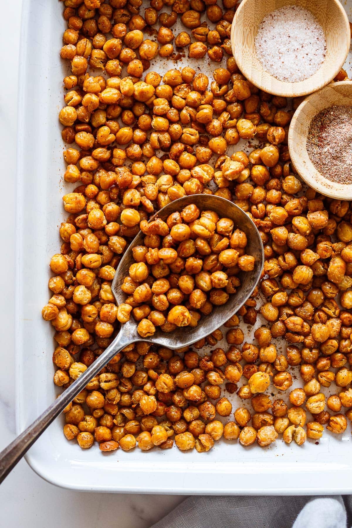 baking sheet with roasted chickpeas with two small bowls of salt and seasonings, with silver spoon scooping some of the chickpeas up.