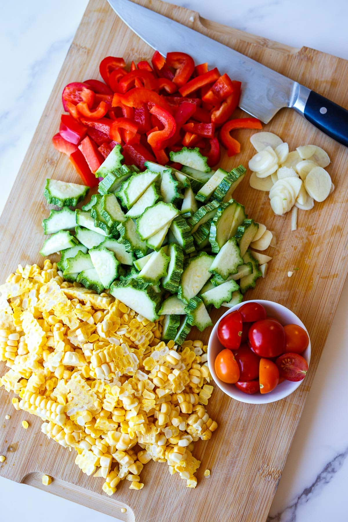 wood cutting board with corn, zucchini, red bell pepper, garlic, and cherry tomatoes next to sharp knife.