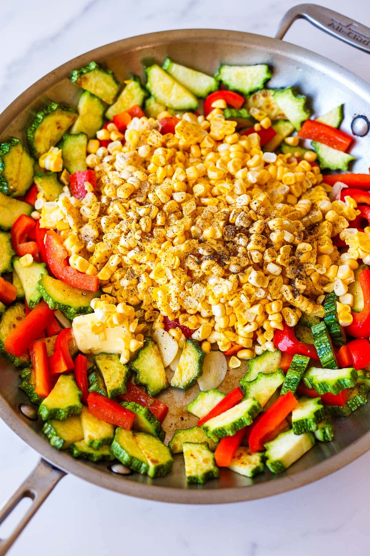 sauteed zucchini, peppers, and garlic, with pile of fresh corn added to skillet with a pat of butter.