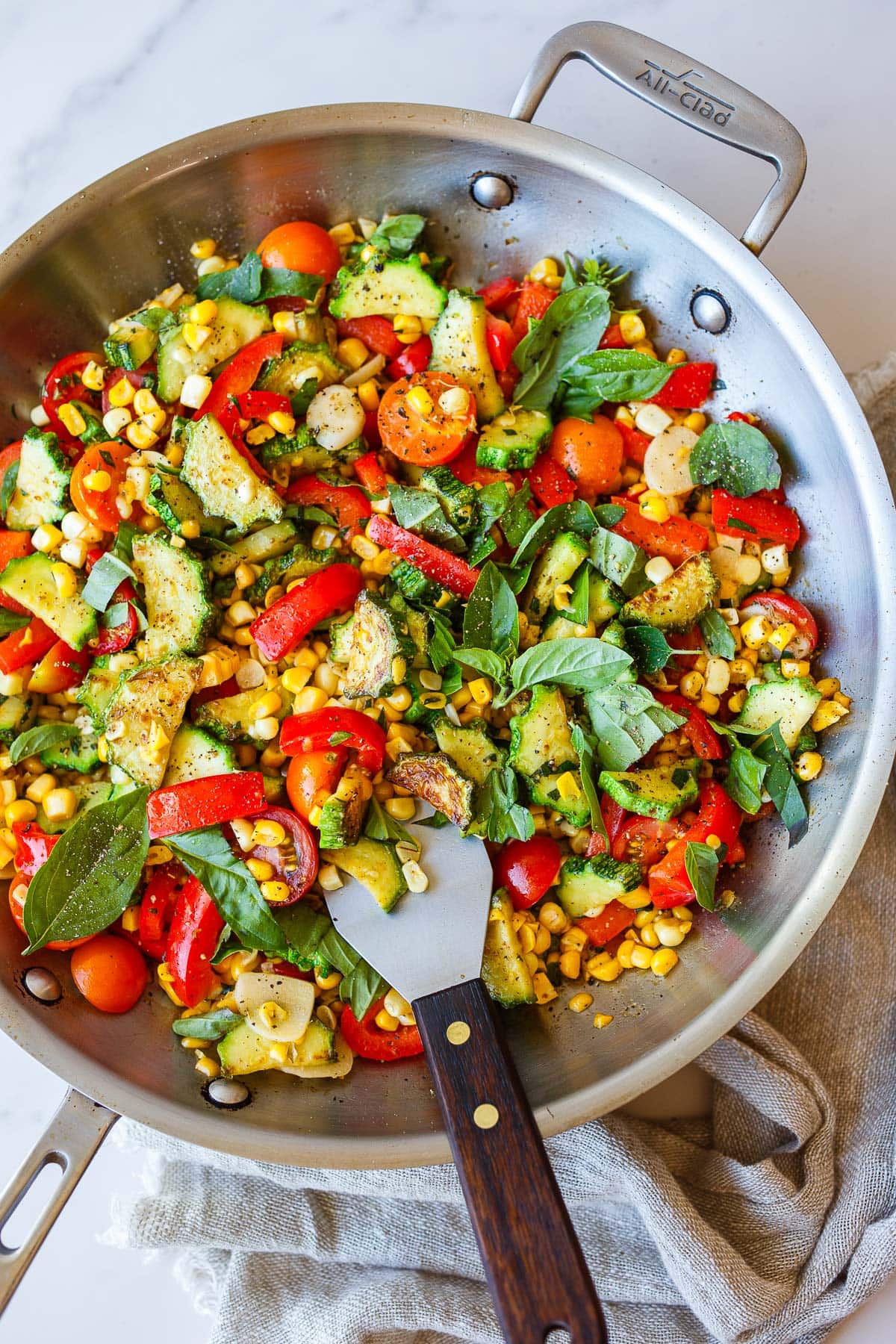 sautéed zucchini with bell peppers, corn, cherry tomatoes, and basil in large stainless steel skillet with metal spatula.