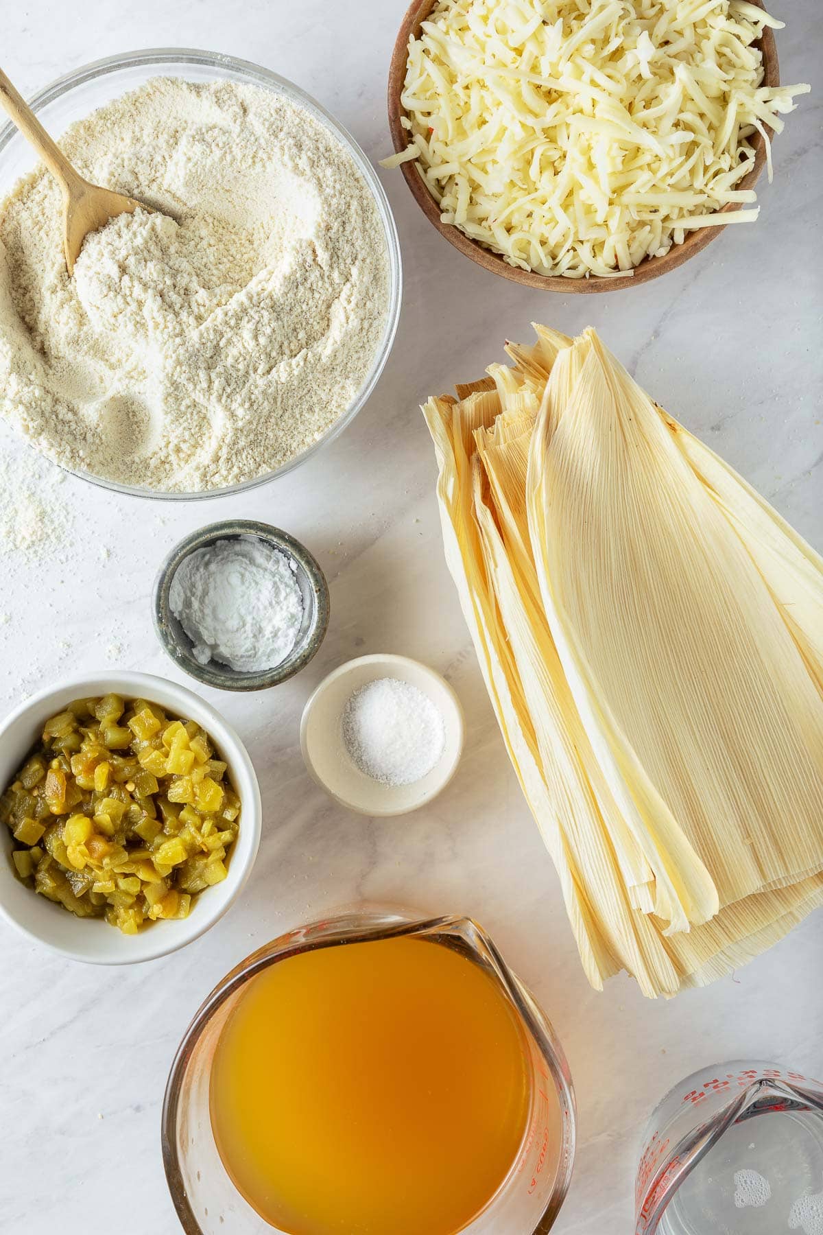 ingredients for tamales - bowl of grated cheese, bowl of masa harina with wood spoon, corn husks, small bowl of baking powder, salt, and green chilies, large measuring cup of broth, and a measuring cup of melted coconut oil. 