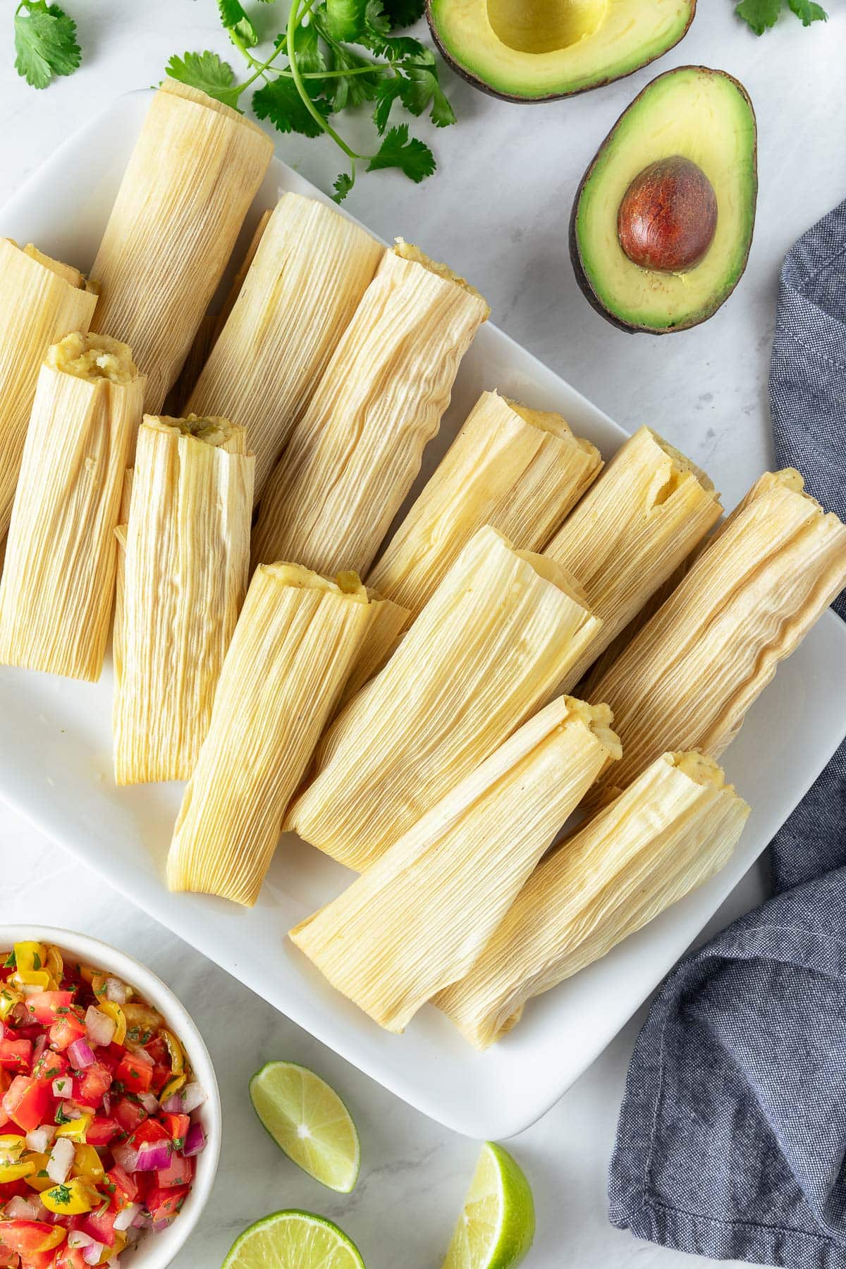 tamales on a rectangular serving platter, surrounded by avocado, cilantro, limes, and pico de gallo. 