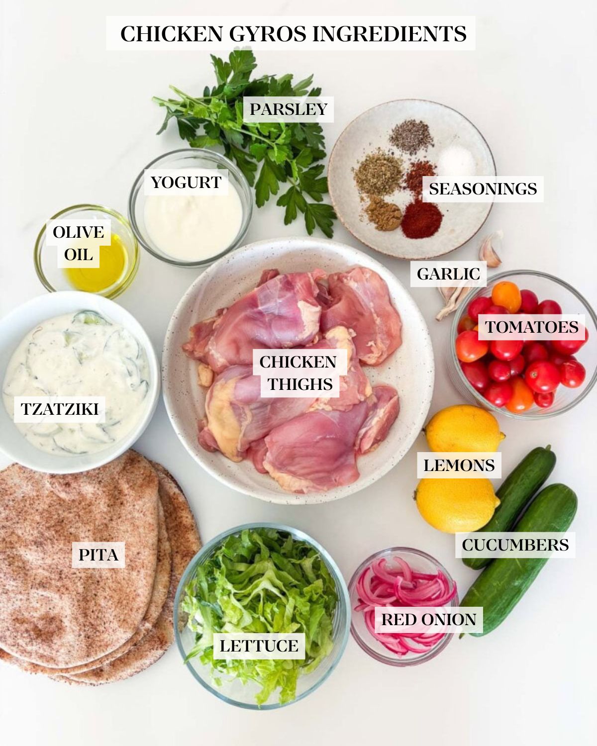 chicken gyros ingredients labeled and arranged in bowls - parsley, seasonings, garlic, tomatoes, lemons, cucumbers, red onion, lettuce, pita, tzatziki, olive oil, yogurt, and chicken thighs. 