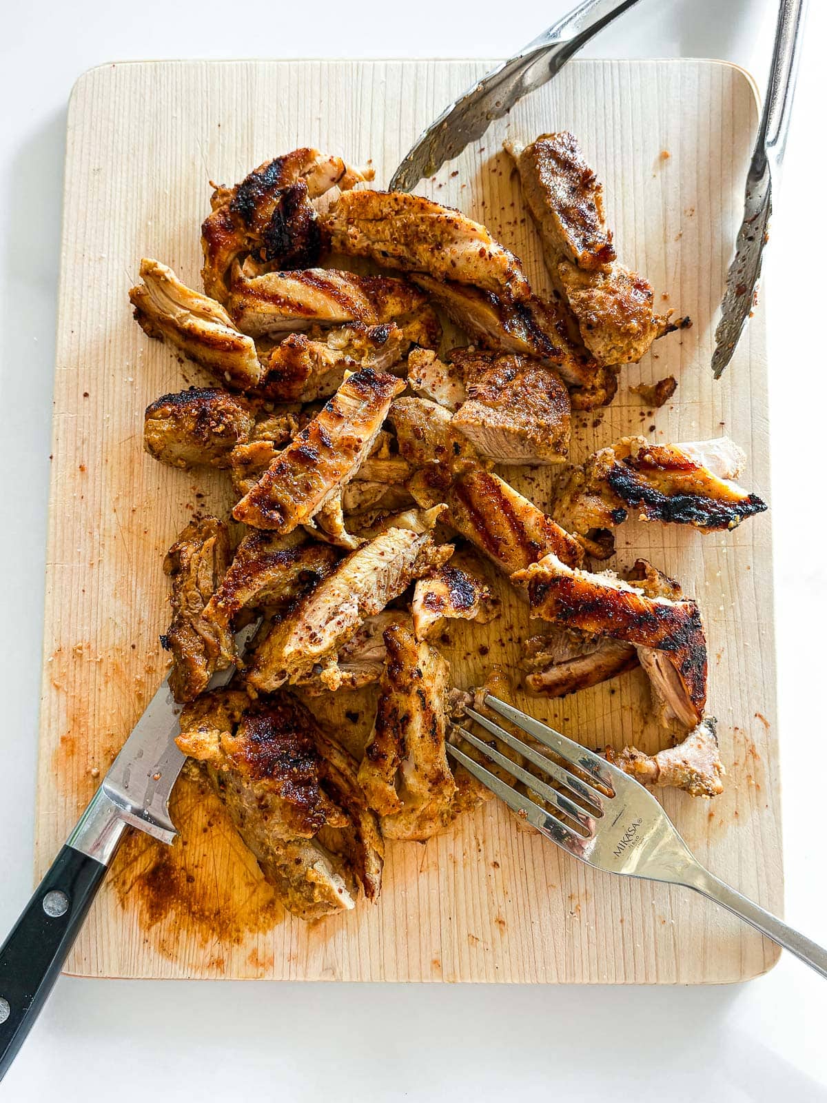 wood board with torn chicken thighs with grill marks, next to tongs, fork, and knife. 