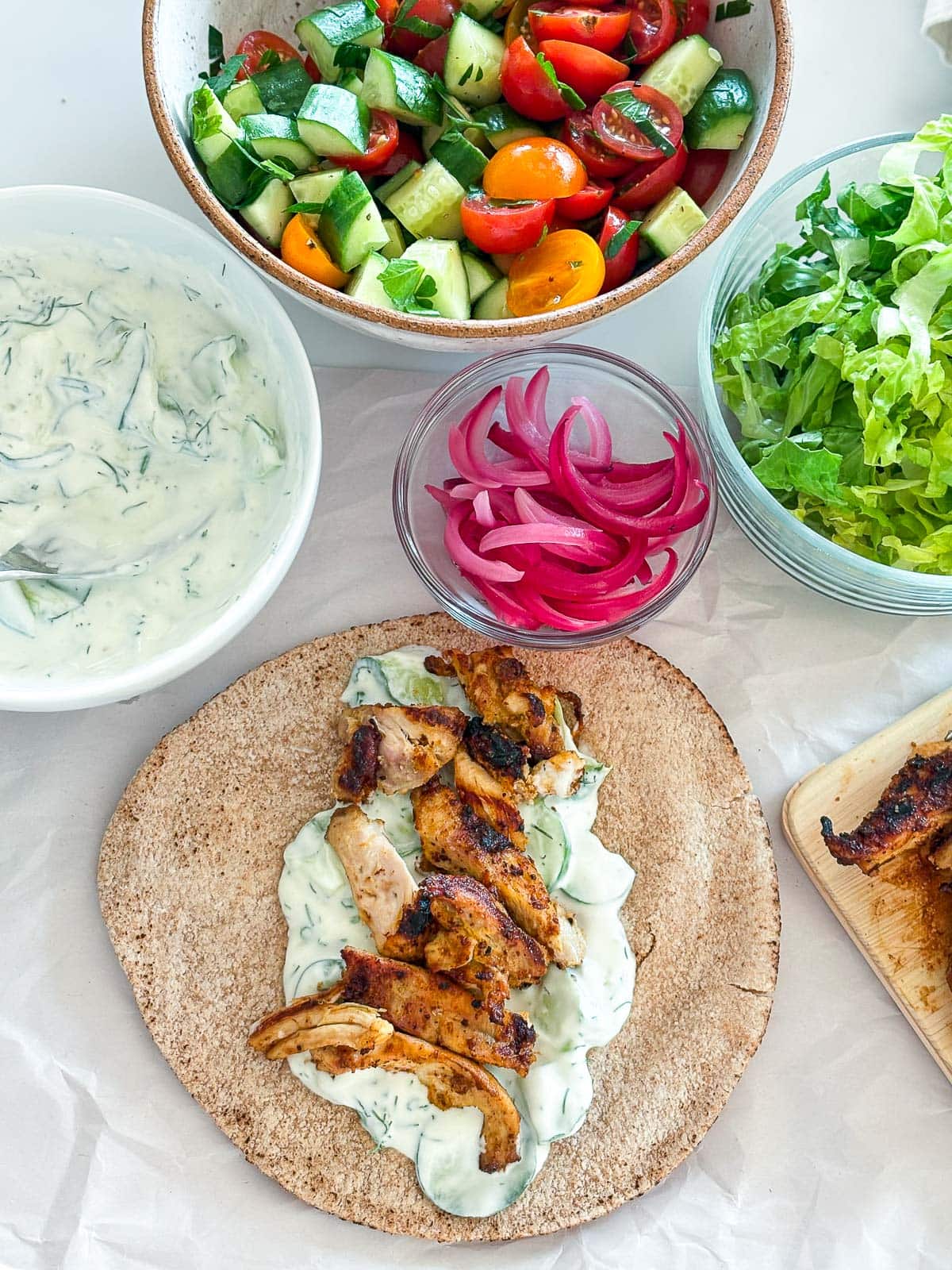 assembling chicken gyros - pita with tzatziki sauce with marinated grilled chicken pieces on top - next to bowls of tzatziki sauce, tomato cucumber salad, pickled onions, and shredded lettuce. 